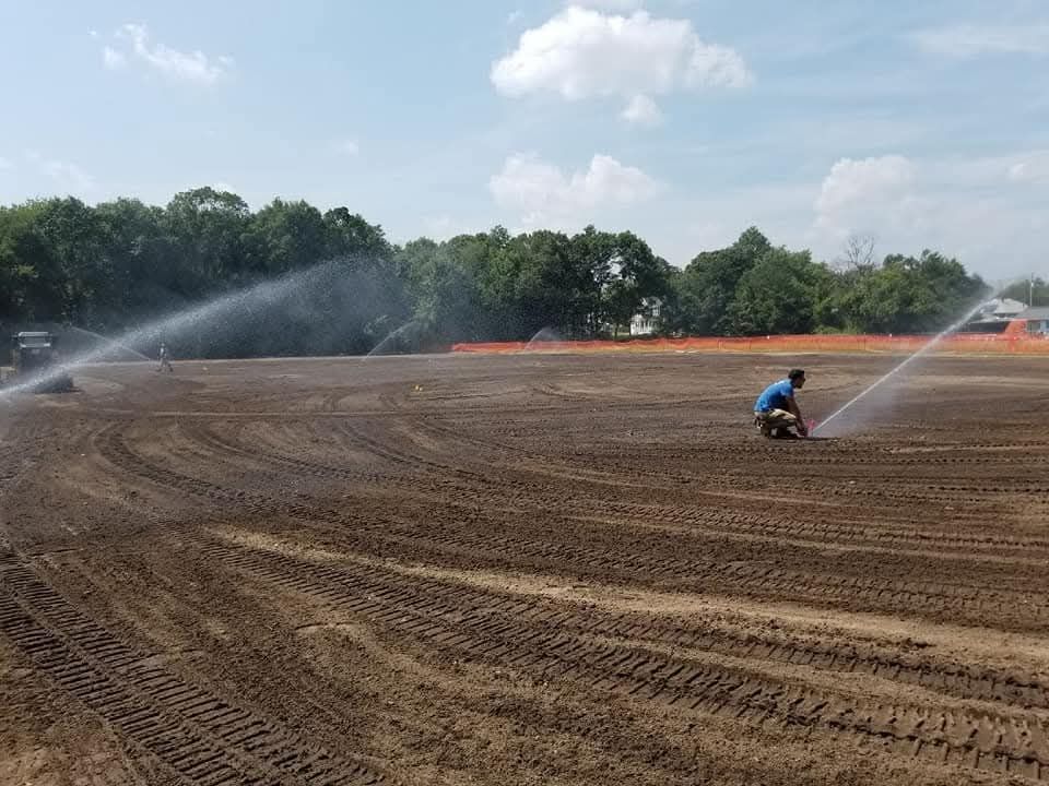 A man is kneeling in a field with a sprinkler spraying water