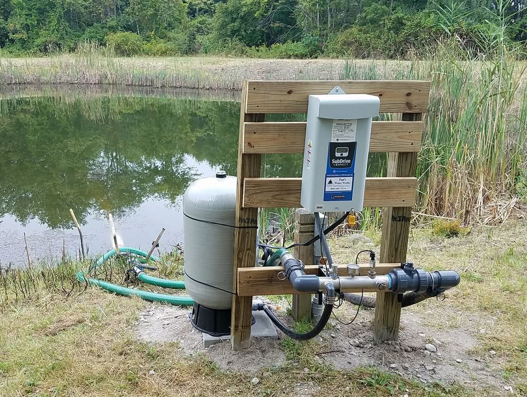 A water pump is sitting on a wooden post next to a lake.
