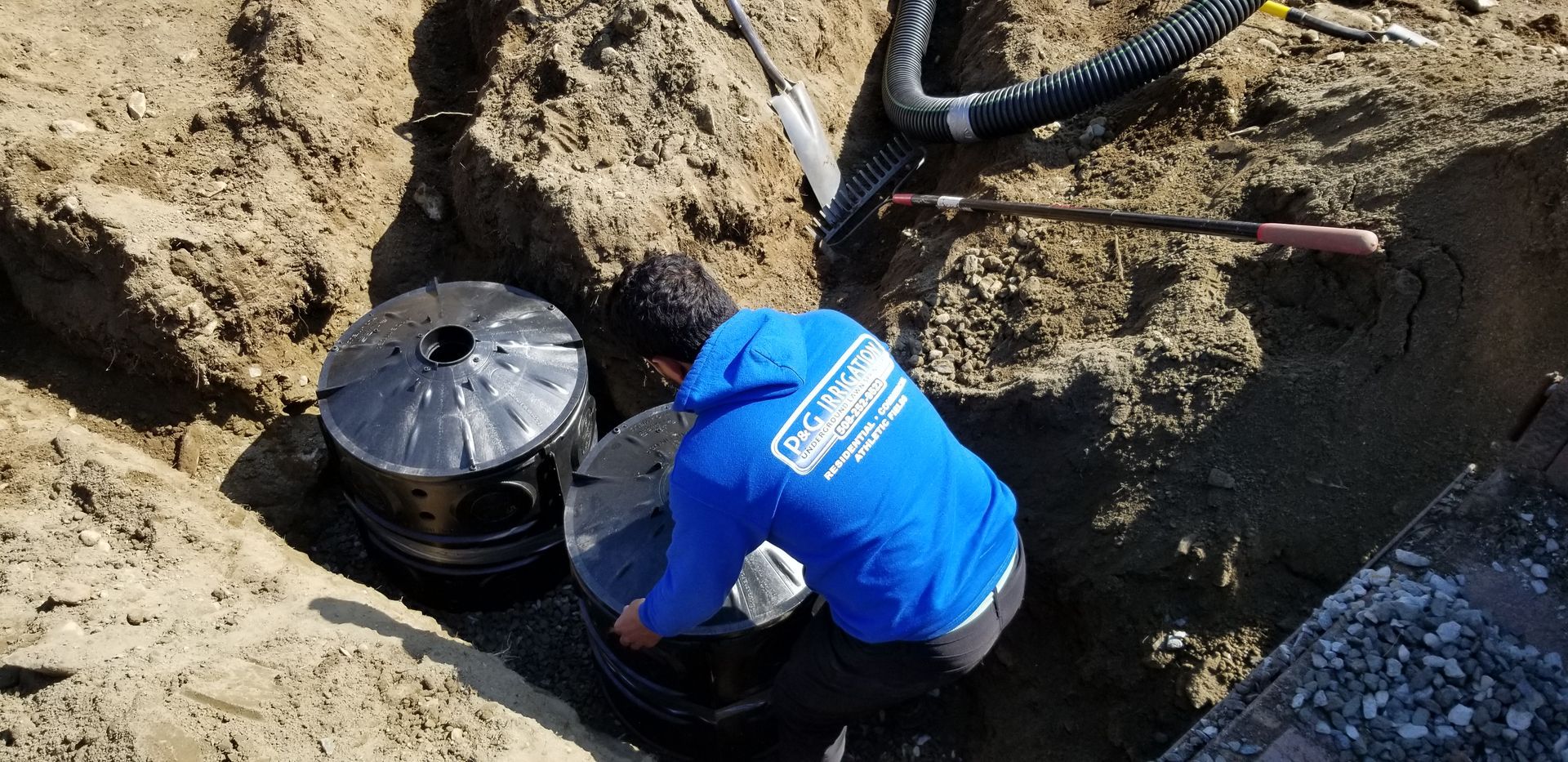 A man in a blue hoodie is working on a septic tank in the dirt.