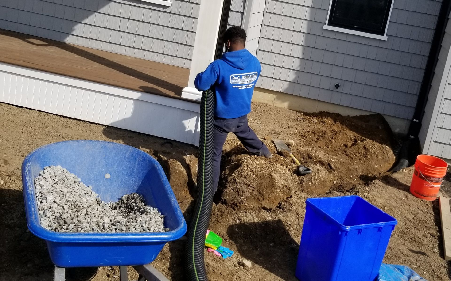 A man is kneeling in the dirt next to a wheelbarrow filled with dirt.