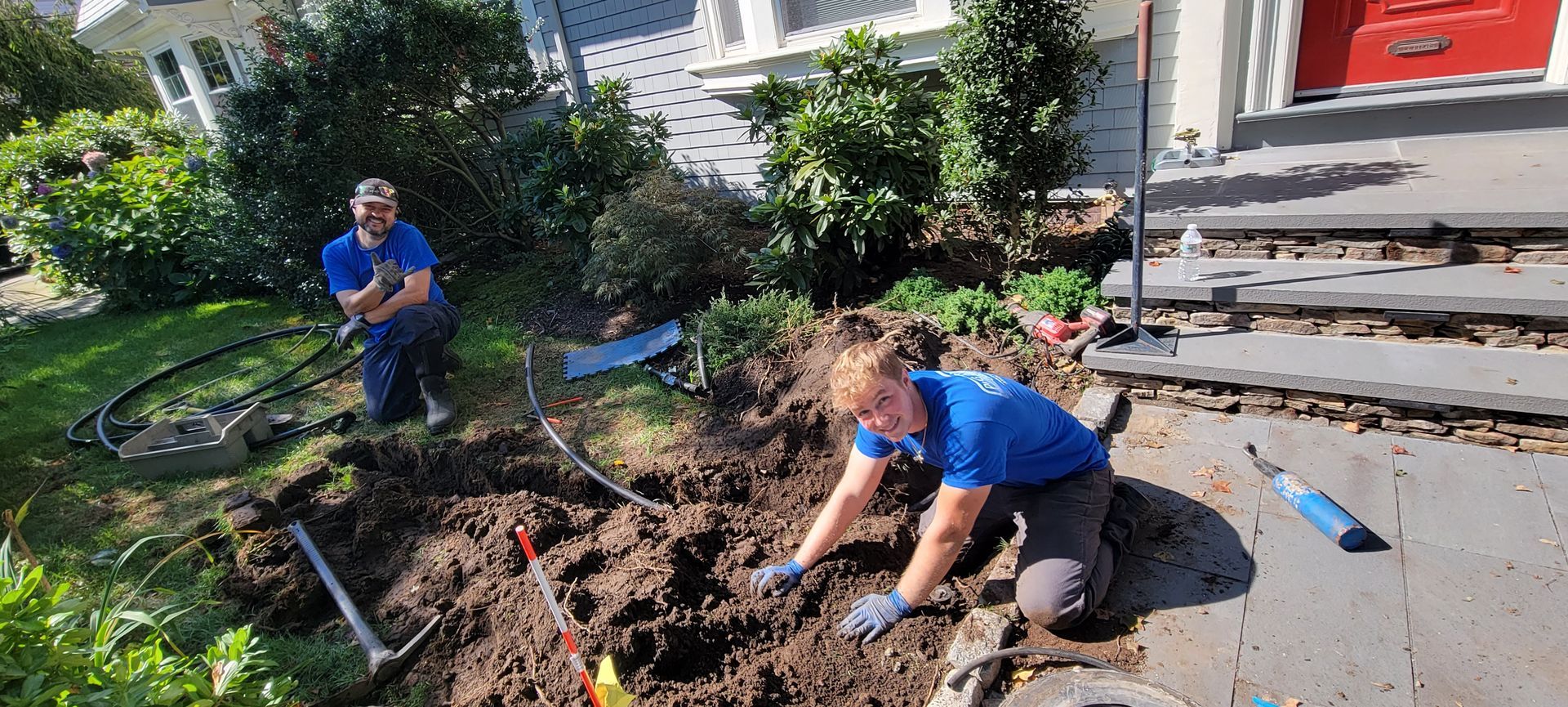 Two men are digging in the dirt in front of a house.