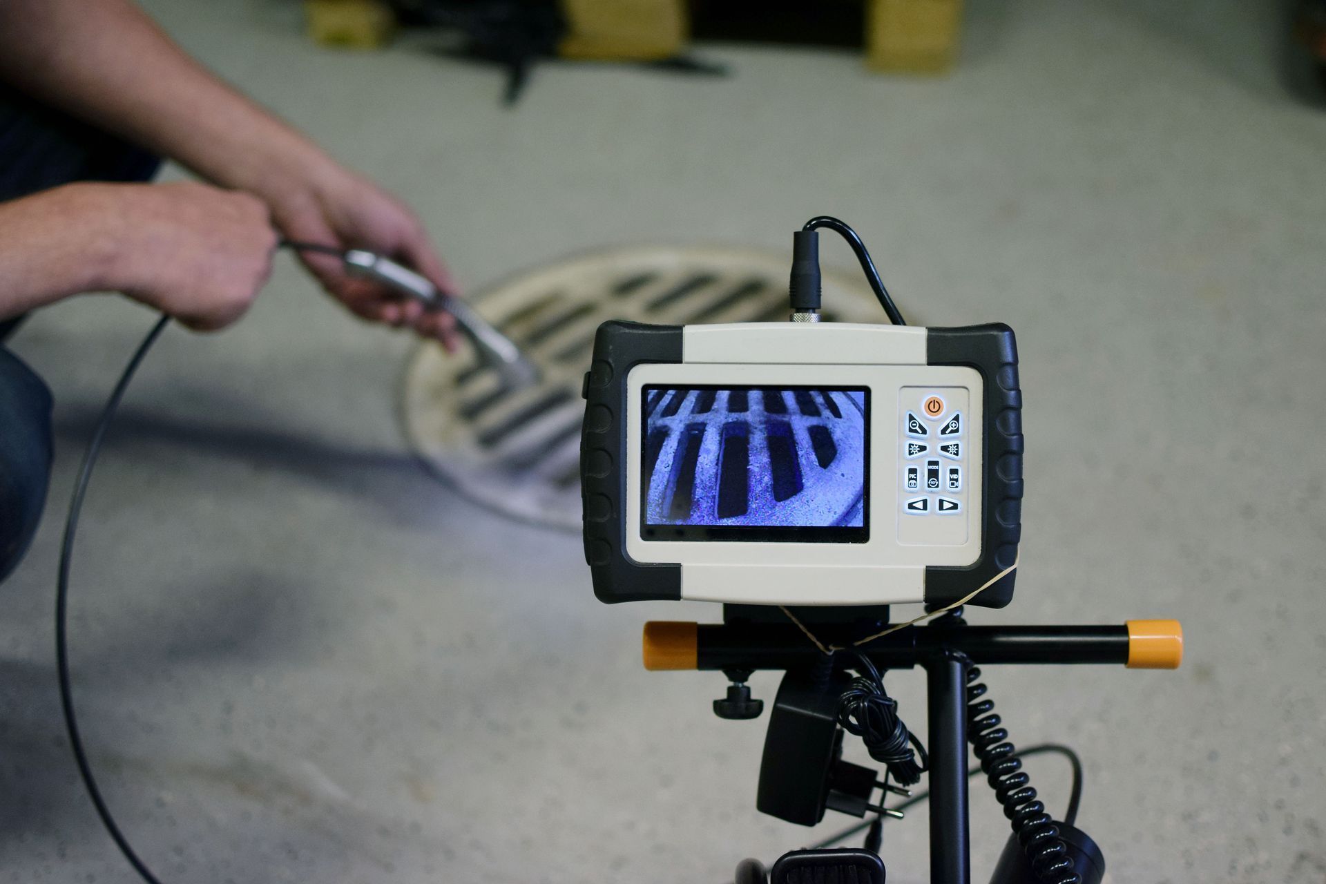 A camera on a tripod displaying a drain's interior with a technician holding a probe. Indoor setting.
