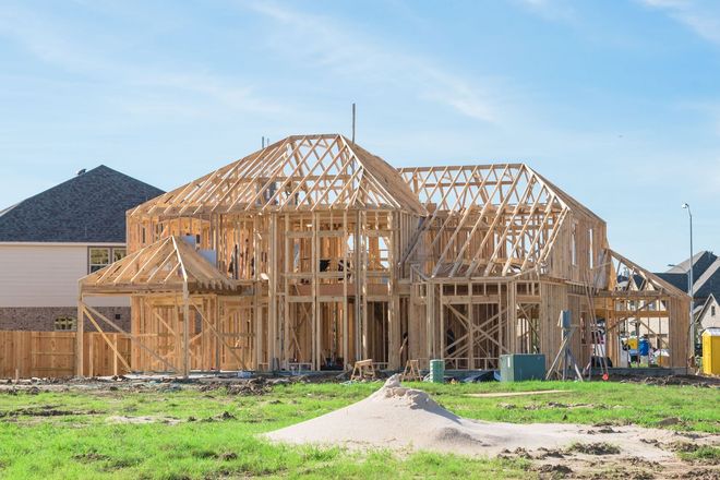 Wooden frame of a house under construction on a grassy lot, with nearby houses and blue sky.