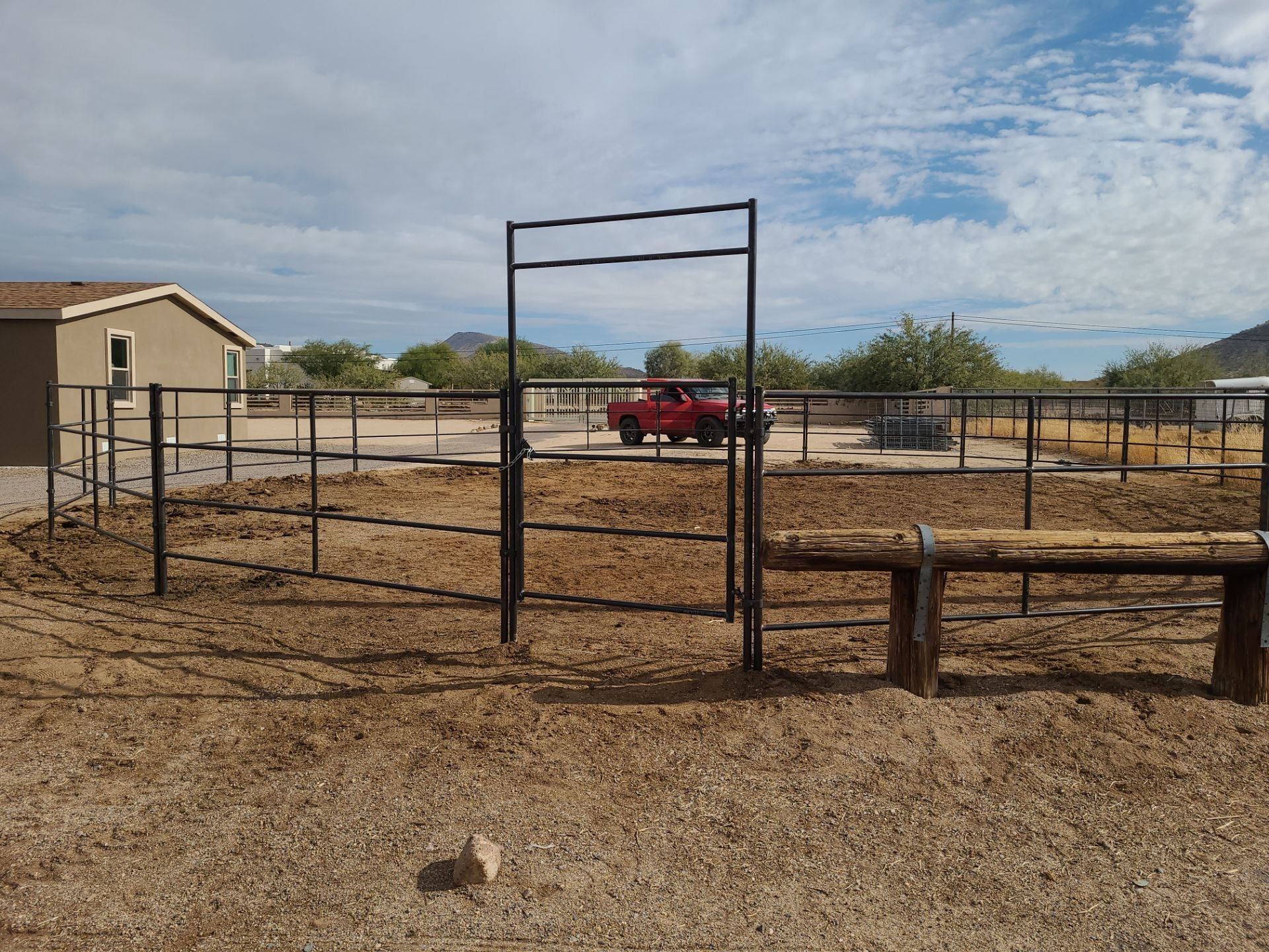 Metal pen with gate, empty dirt ground, small house, red truck in background under partly cloudy sky.