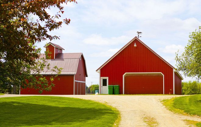 Two red barns on a grassy hill; a gravel driveway leading to the open garage door.