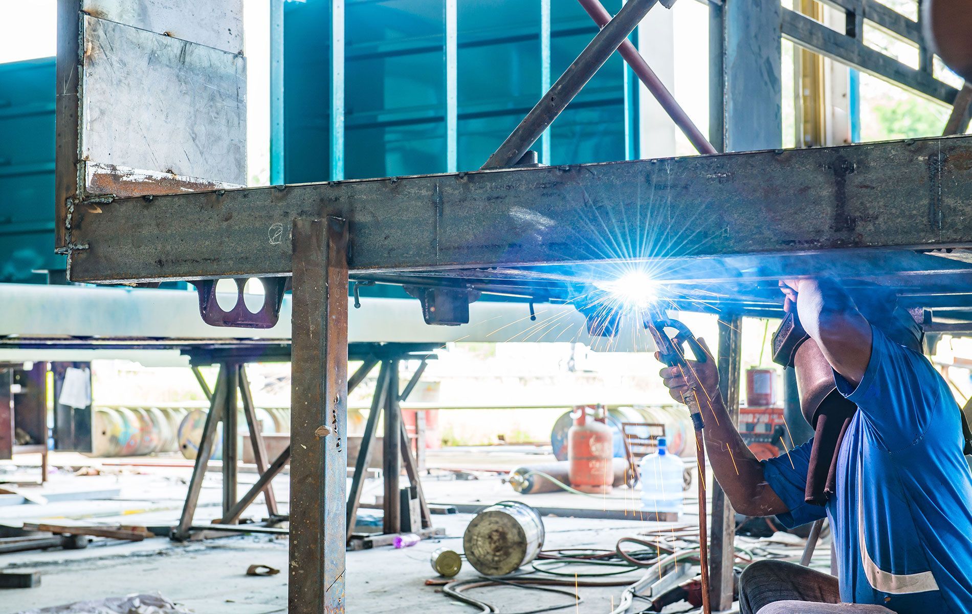 Welder in blue uniform using a welding torch to join metal beams in a workshop.