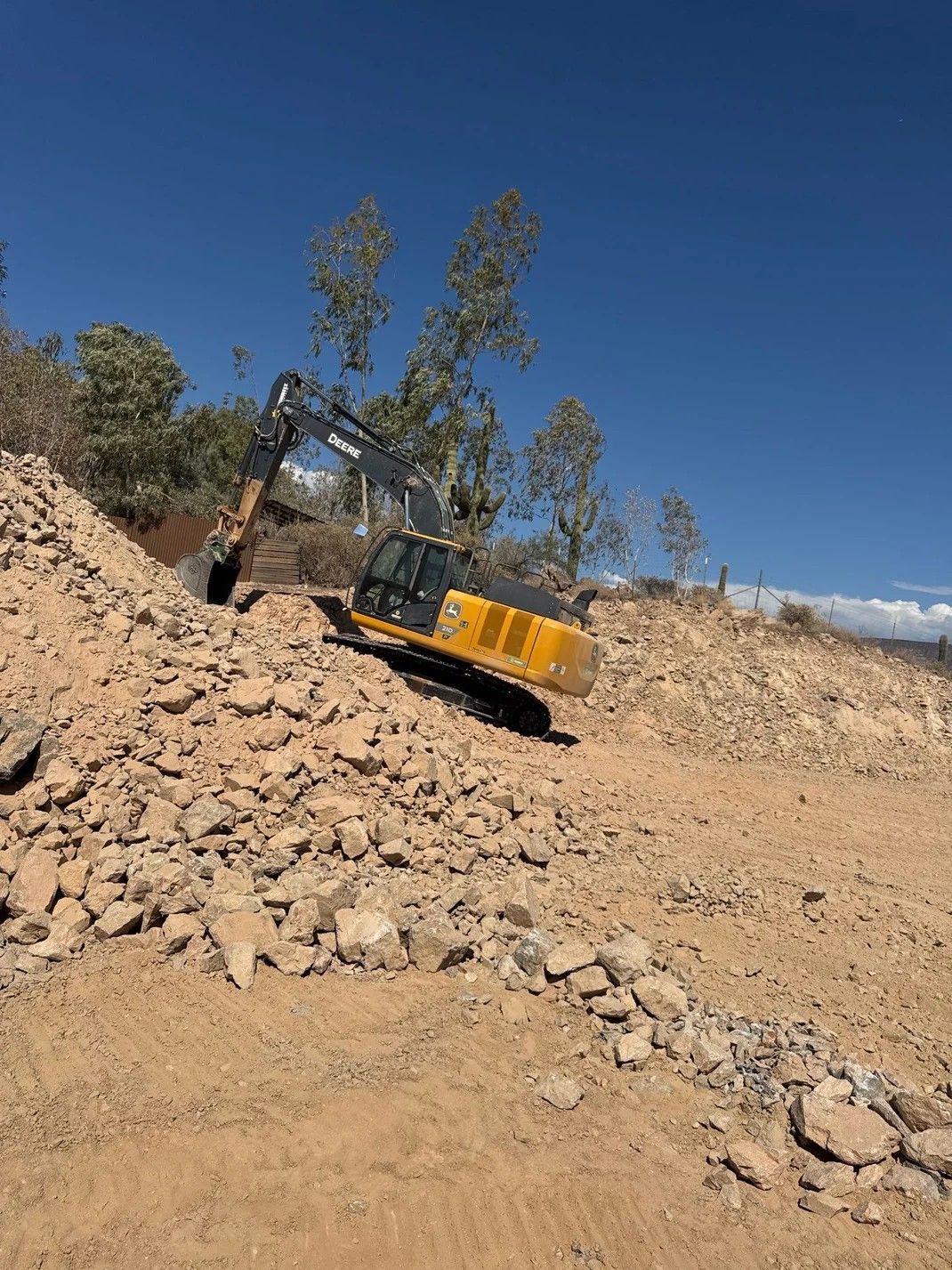 Excavator on a rocky hillside under a blue sky, digging into a dirt and rock pile.