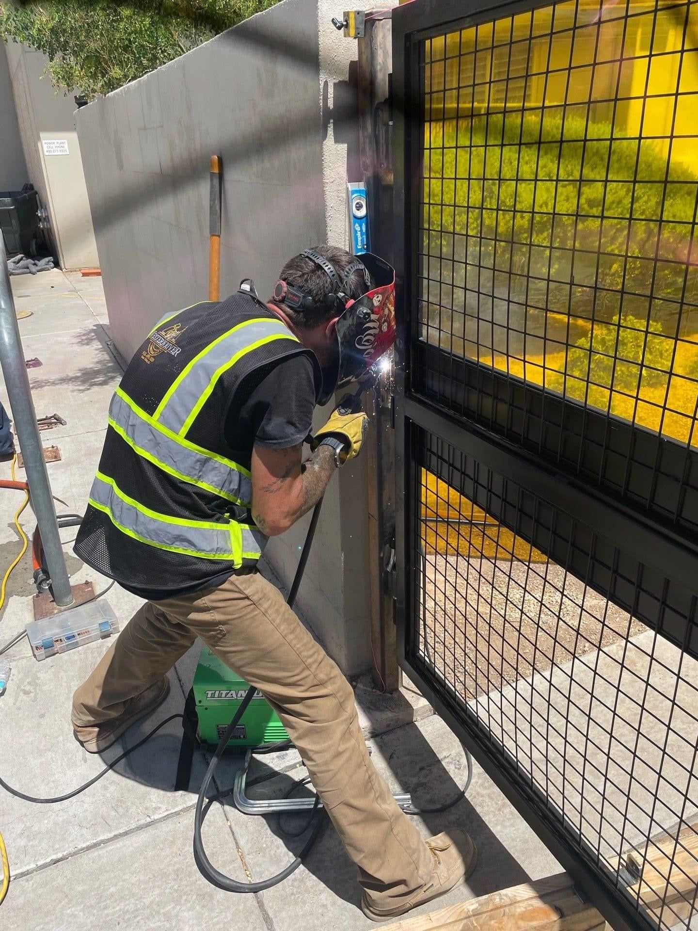 Person welding metal gate outdoors, wearing safety vest, brown pants, and gloves.