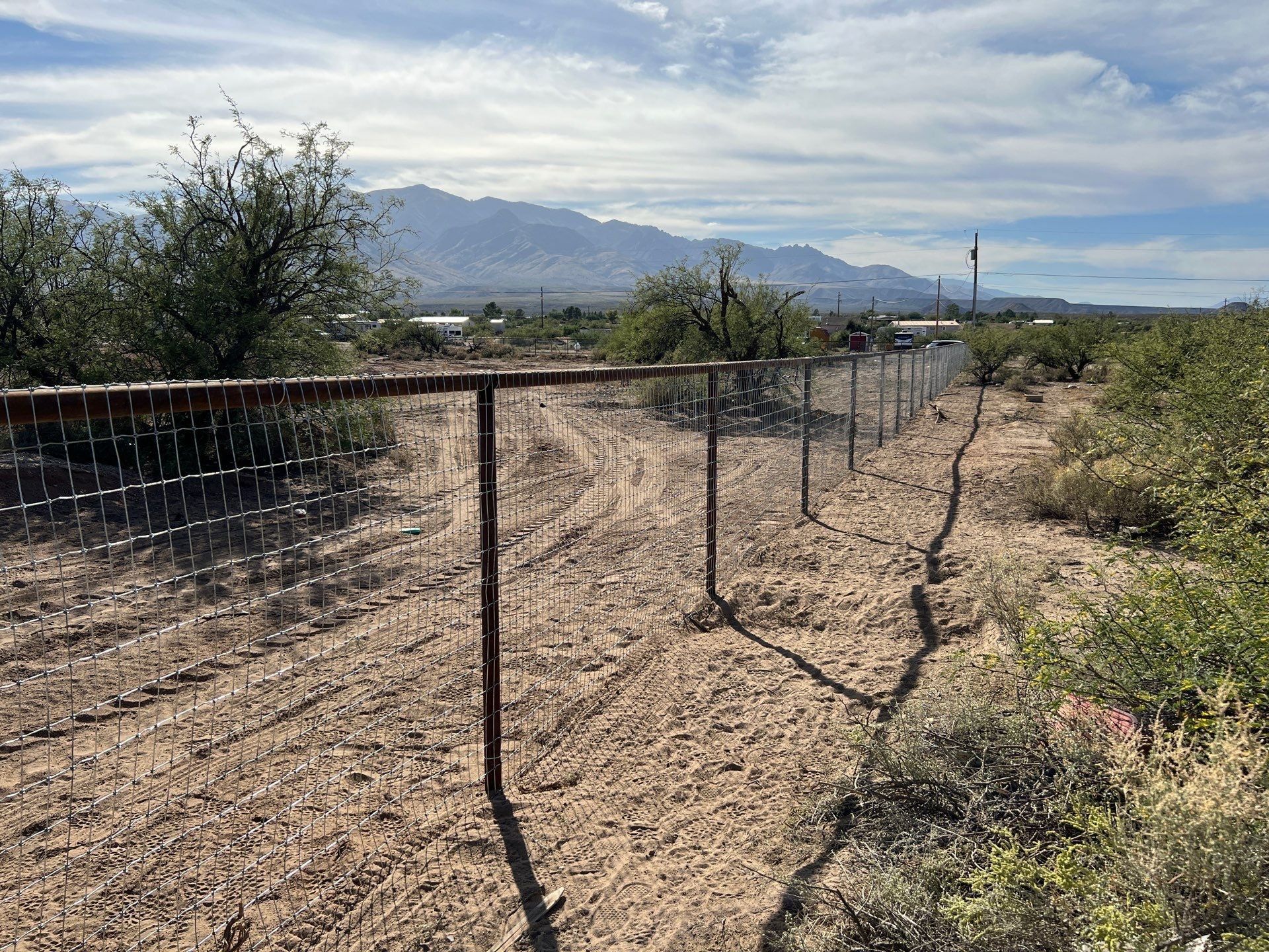 Chain-link fence in a desert landscape; mountains and vegetation in the background under a blue sky.