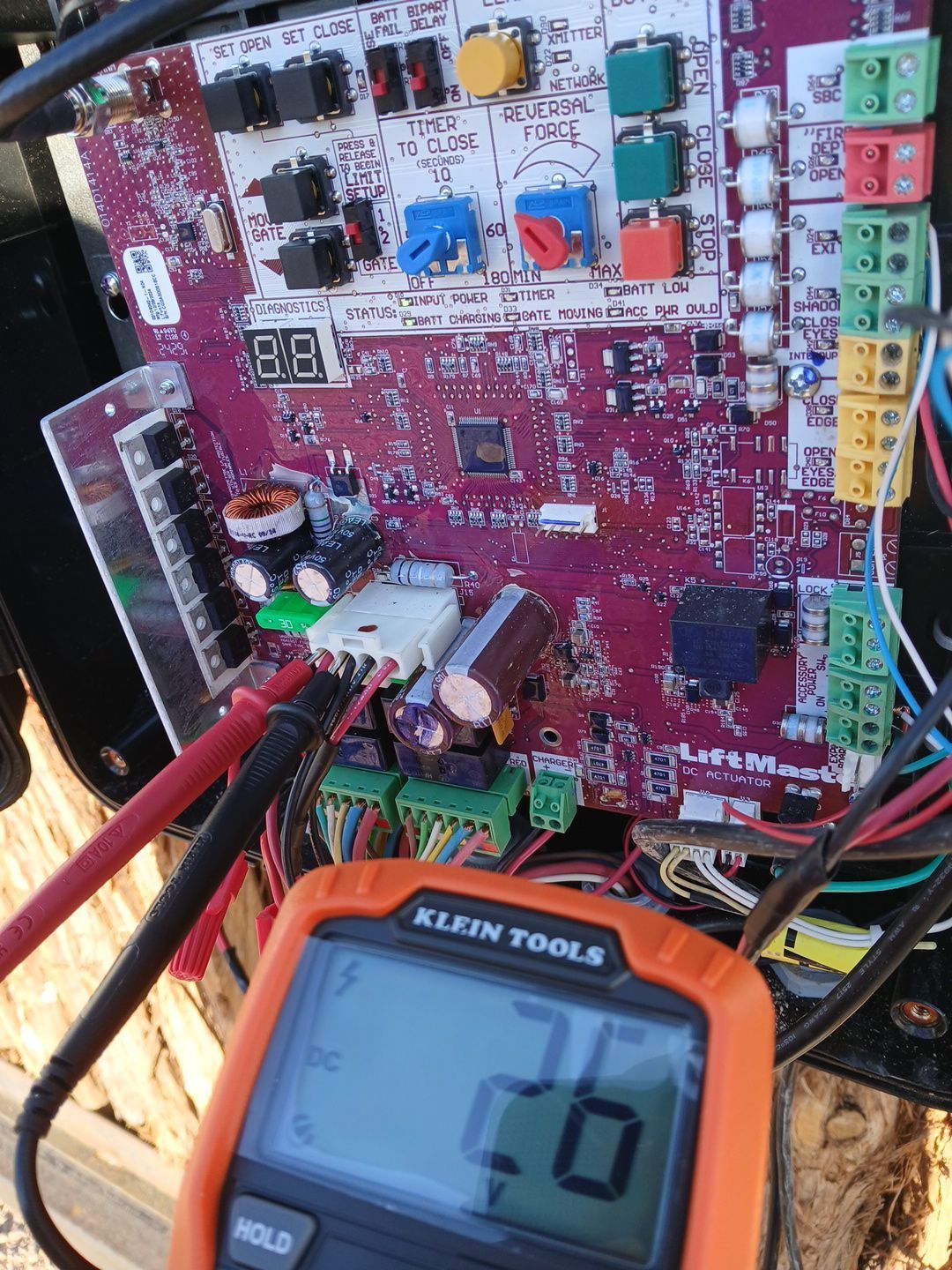 A multimeter testing a circuit board with multicolored terminals and components. Digital display reads 27 volts DC.