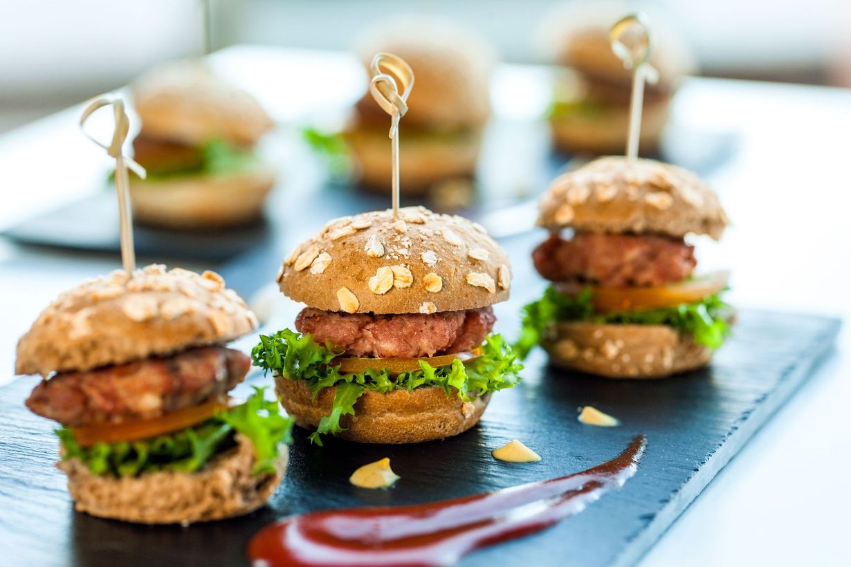 Mini burgers on slate platter, garnished with lettuce and sauce, with blurred appetizers in the background