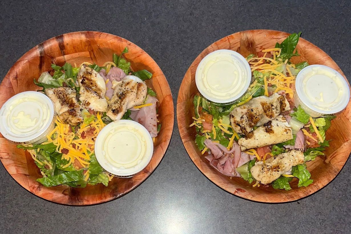 Two salads in wooden bowls on a dark table, topped with dressing cups and shredded cheese