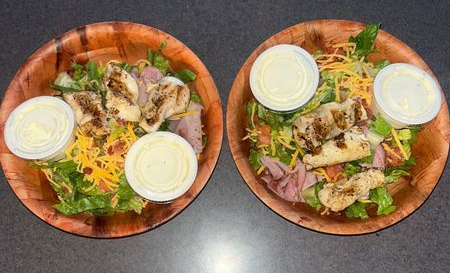 Two wooden bowls of salad with grilled chicken and two dressing cups on a dark table.