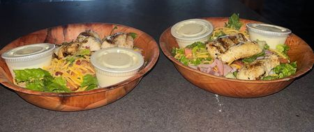Two wooden bowls of grilled fish and salad with dipping sauces on a dark table