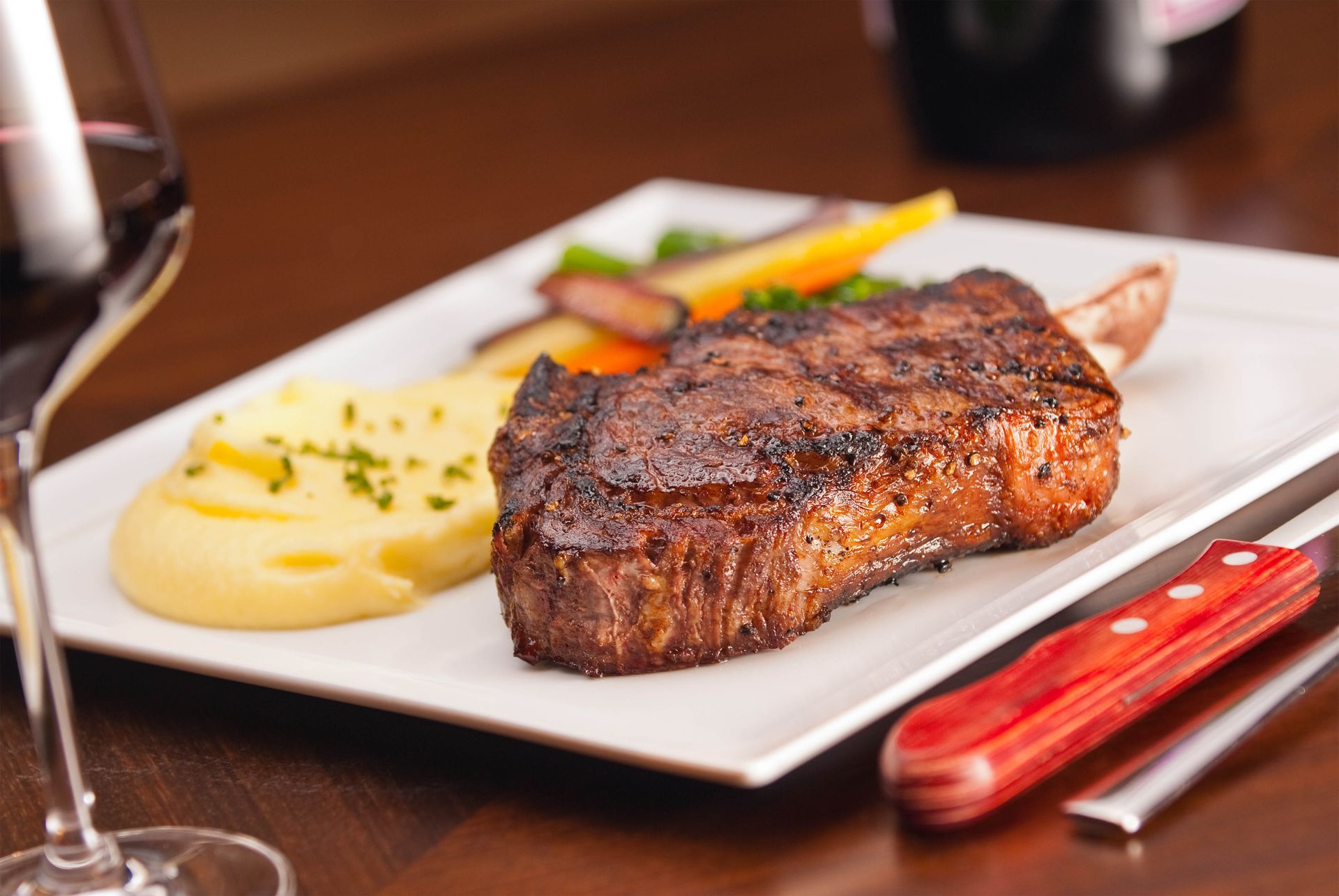 Seared steak on a white plate with mashed potatoes and vegetables on a wooden table, with wine glasses nearby