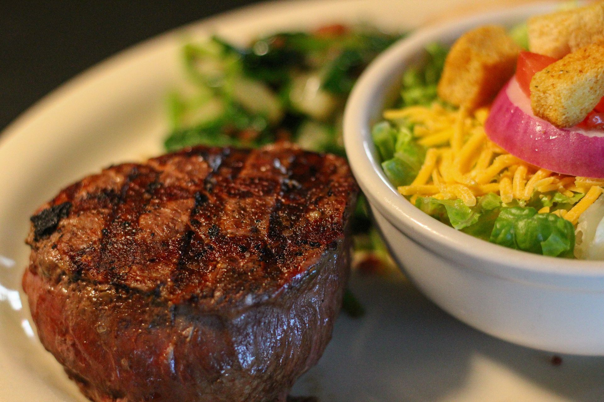 Grilled steak with char marks beside a salad topped with shredded cheese and croutons