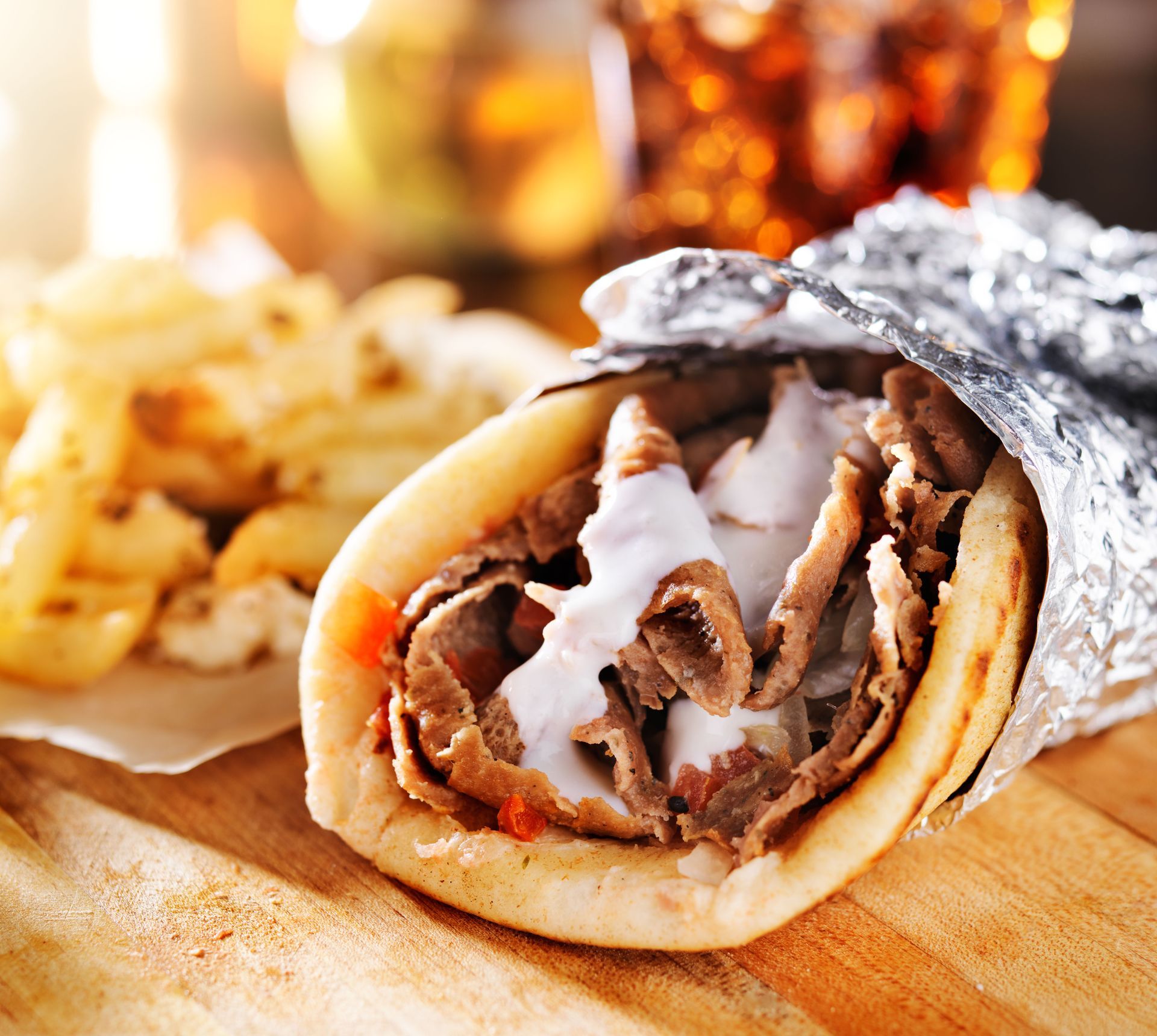 Gyro wrap in foil with meat, sauce, and bread on a wooden board, with chips and drinks in the background