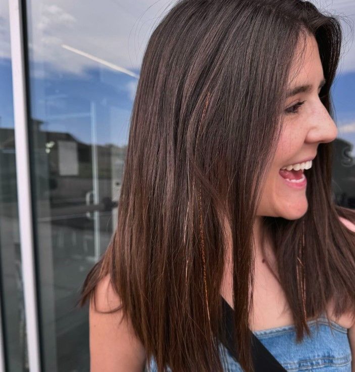 A woman with long brown hair is smiling in front of a window.