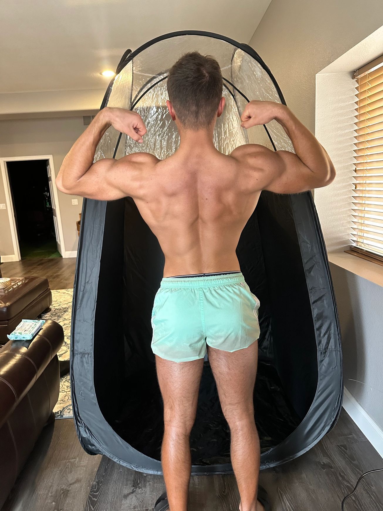 A shirtless man is standing in front of a tanning tent in a living room.