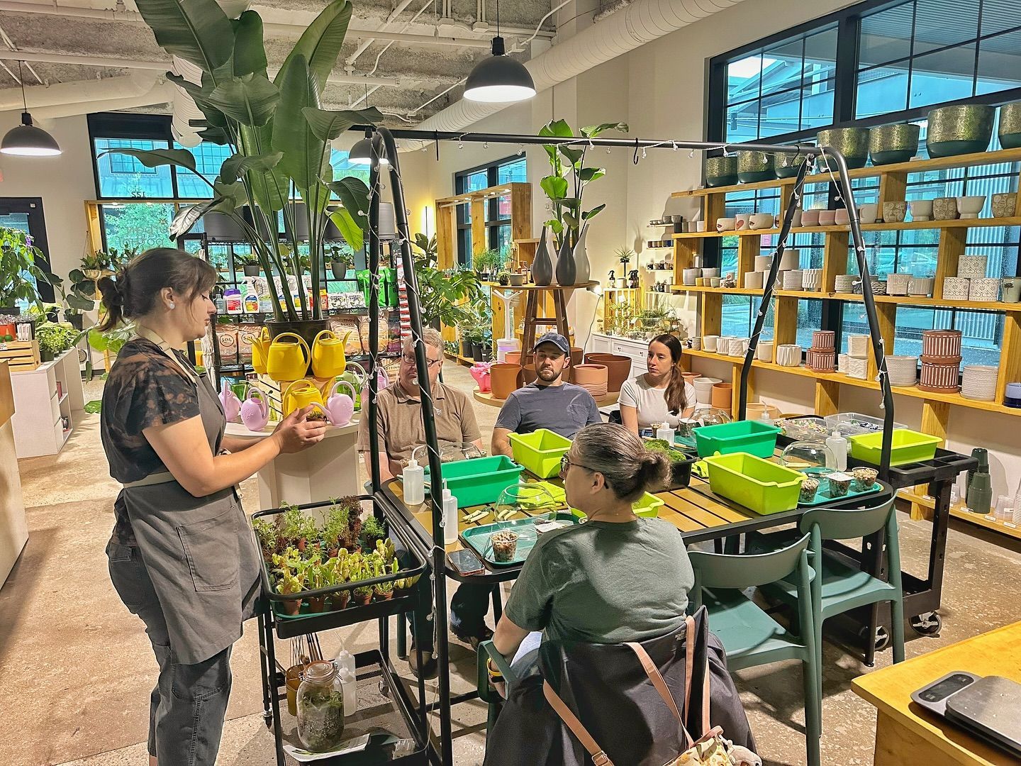 A group of people are sitting around a table in a room filled with potted plants.