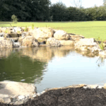 Pond with cascading waterfall, surrounded by rocks and grass, reflecting the sky.