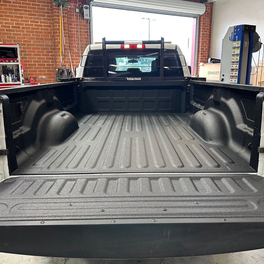 Pickup truck bed with textured black liner, truck bed rack, and open tailgate in a shop.