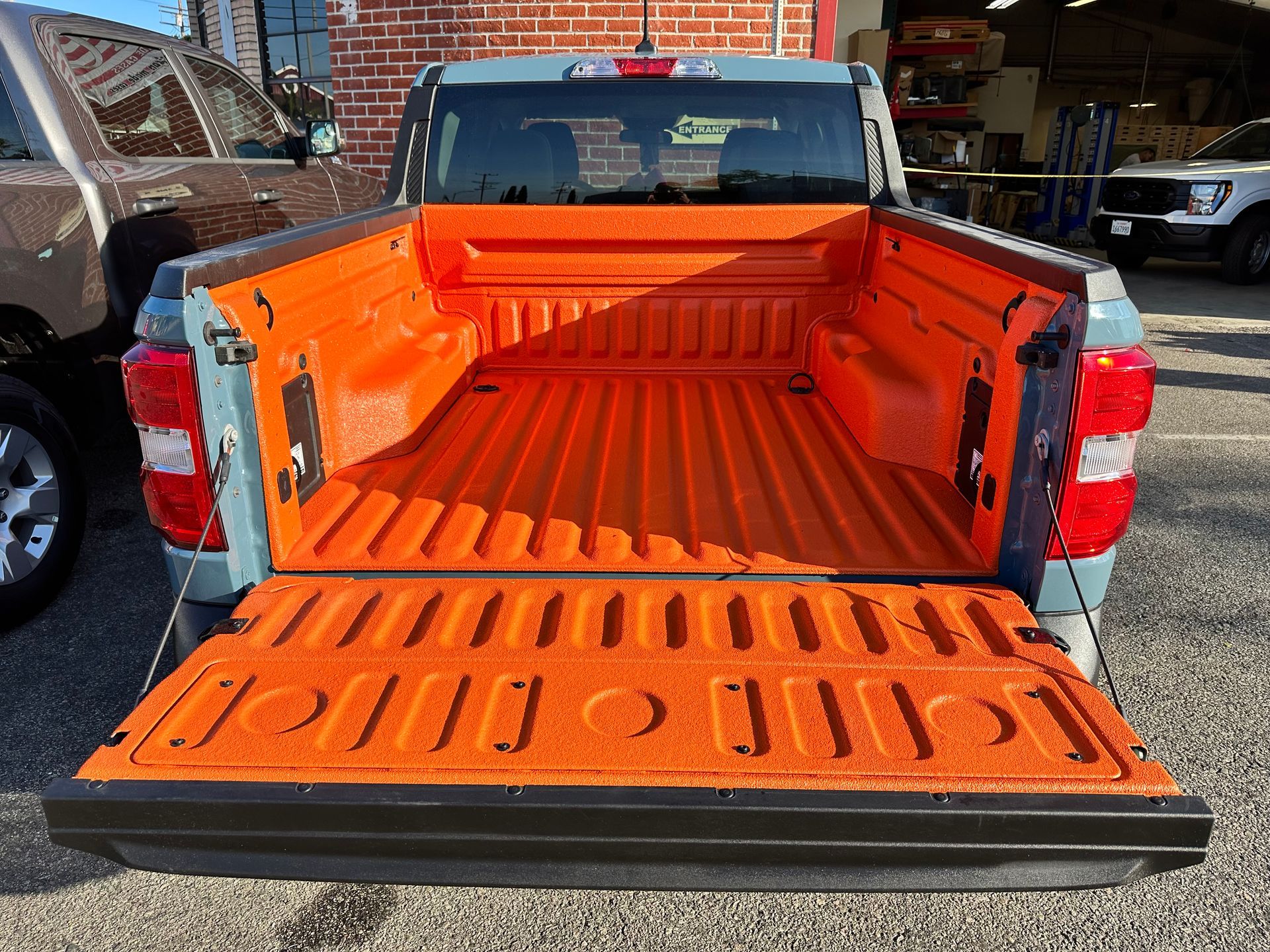 A pickup truck with an open tailgate showing a bright orange spray-in bed liner against a brick building backdrop.