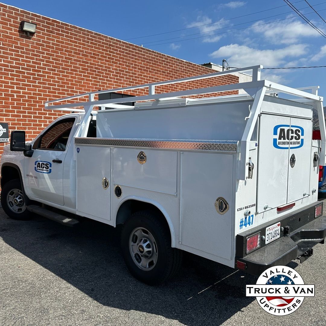 White work truck with utility bed, ladder rack, and ACS logo parked in front of brick wall.