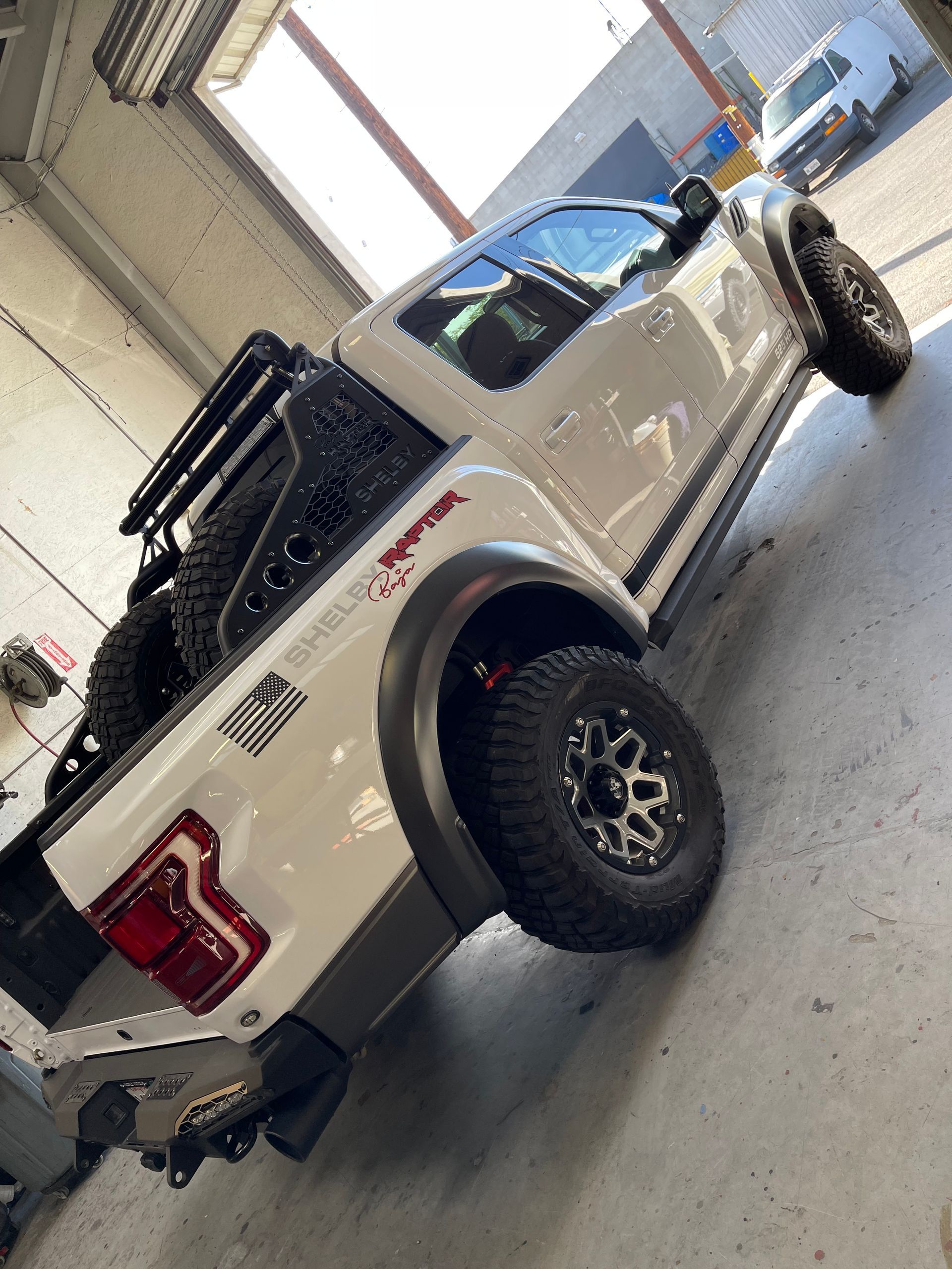 White Shelby Raptor truck with off-road tires and a bed-mounted rack parked in an indoor garage.