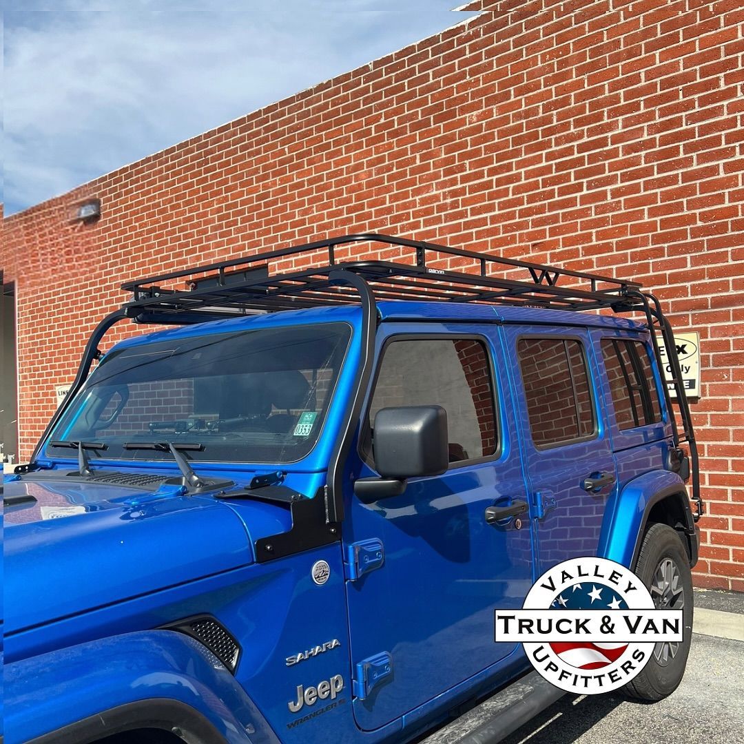 Blue Jeep with black roof rack parked near a brick building.
