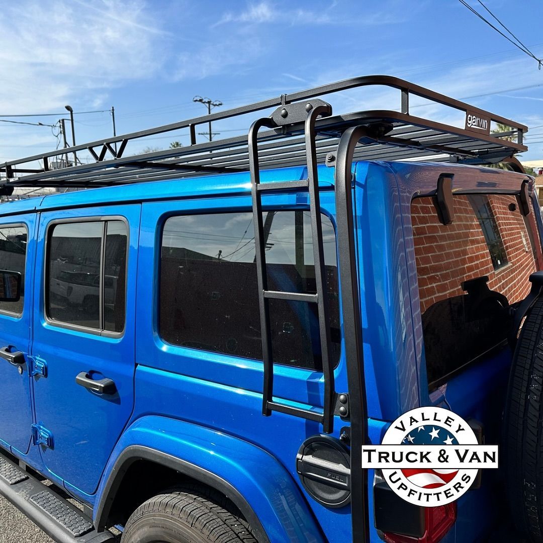 Blue Jeep Wrangler with black roof rack and ladder, under a partly cloudy sky.