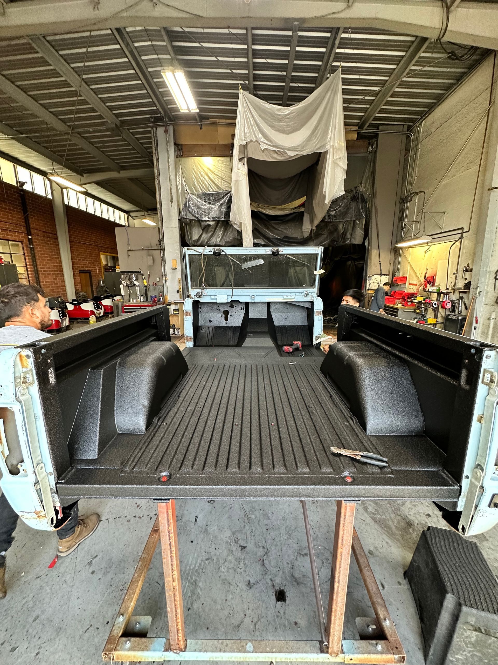 A light blue pickup truck bed with a fresh, black spray-on liner sits on a metal stand inside a brightly lit workshop.