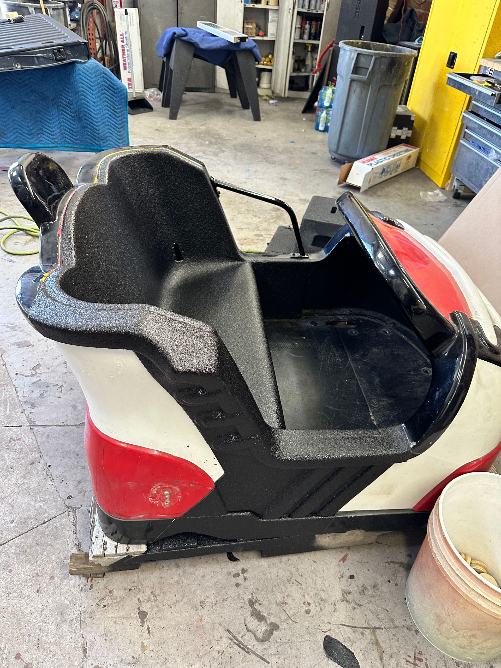 A red and white fiberglass bumper car seat sits on a workshop floor.