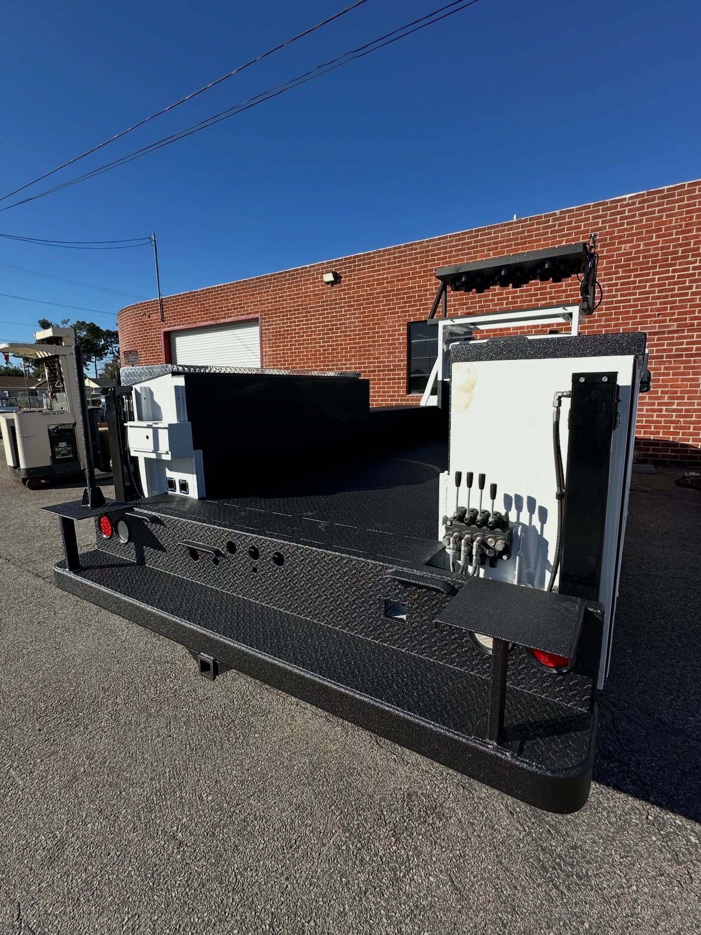 Black and white utility trailer on gravel, with a brick building and blue sky in the background.