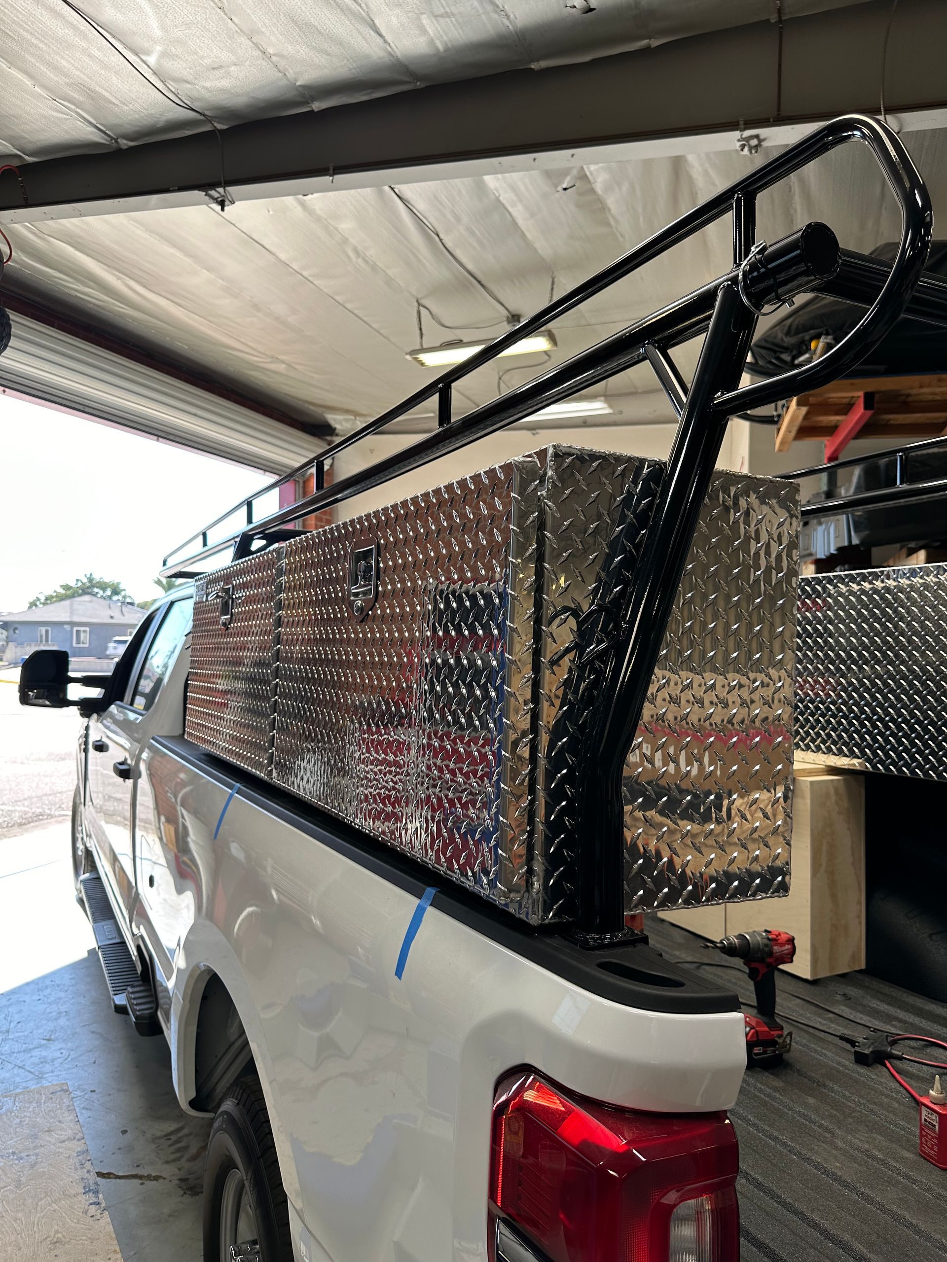 A white pickup truck with a diamond-plate metal tool box and a black overhead utility rack in a garage.