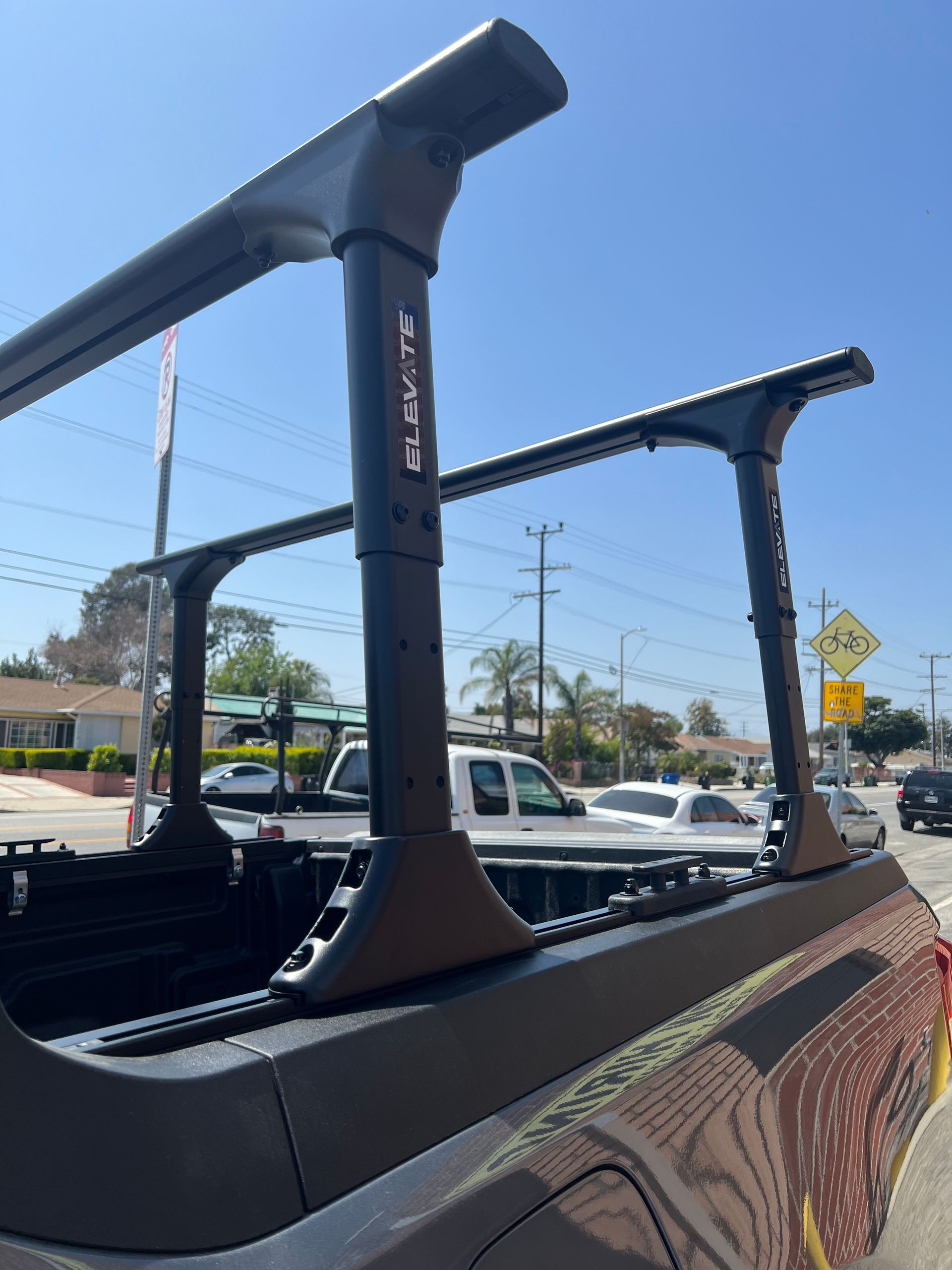 Black adjustable truck bed rack installed on a vehicle, viewed from an outdoor street setting on a sunny day.