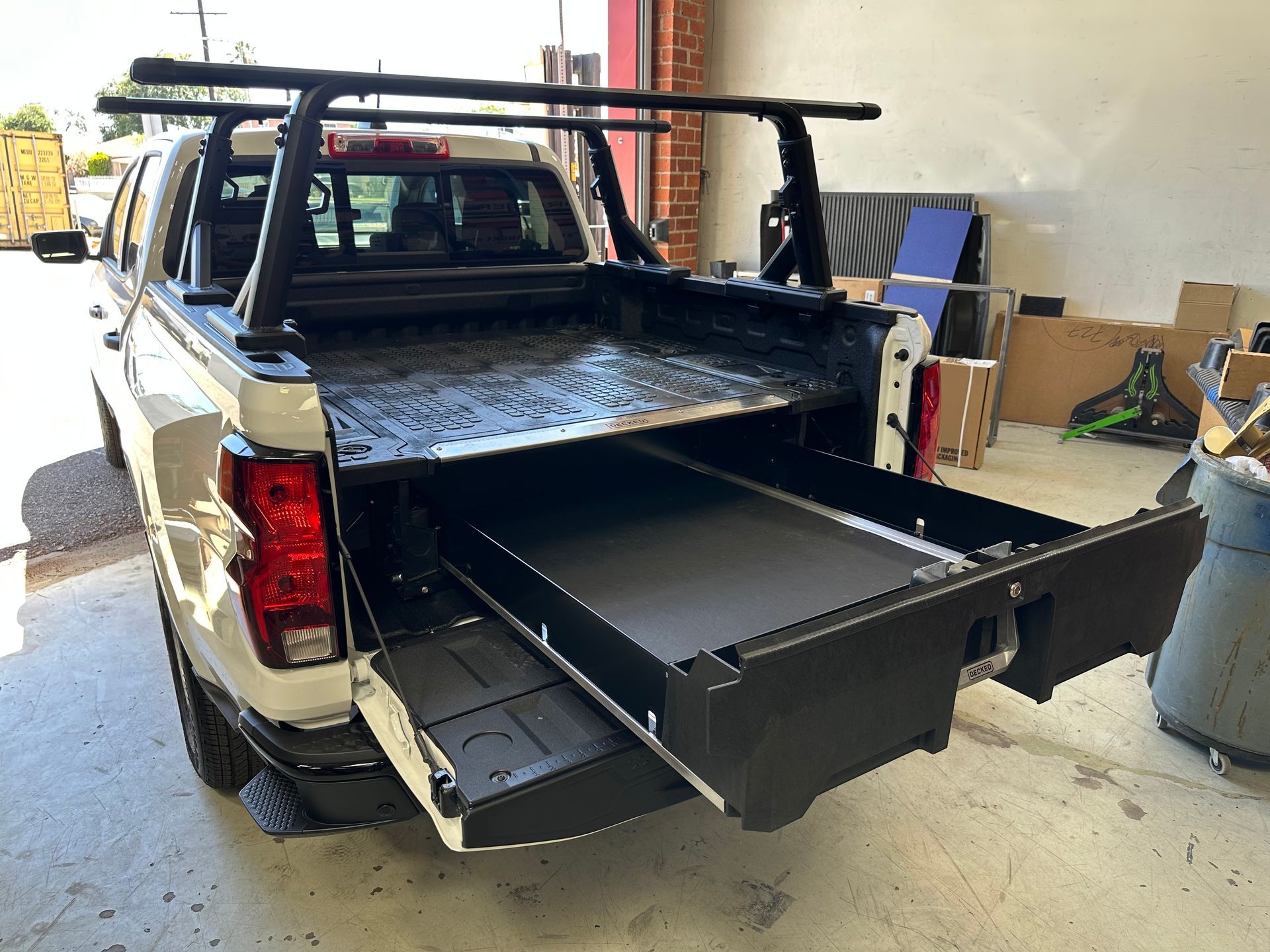 A white pickup truck parked indoors with a black pull-out storage drawer extended from the open cargo bed.