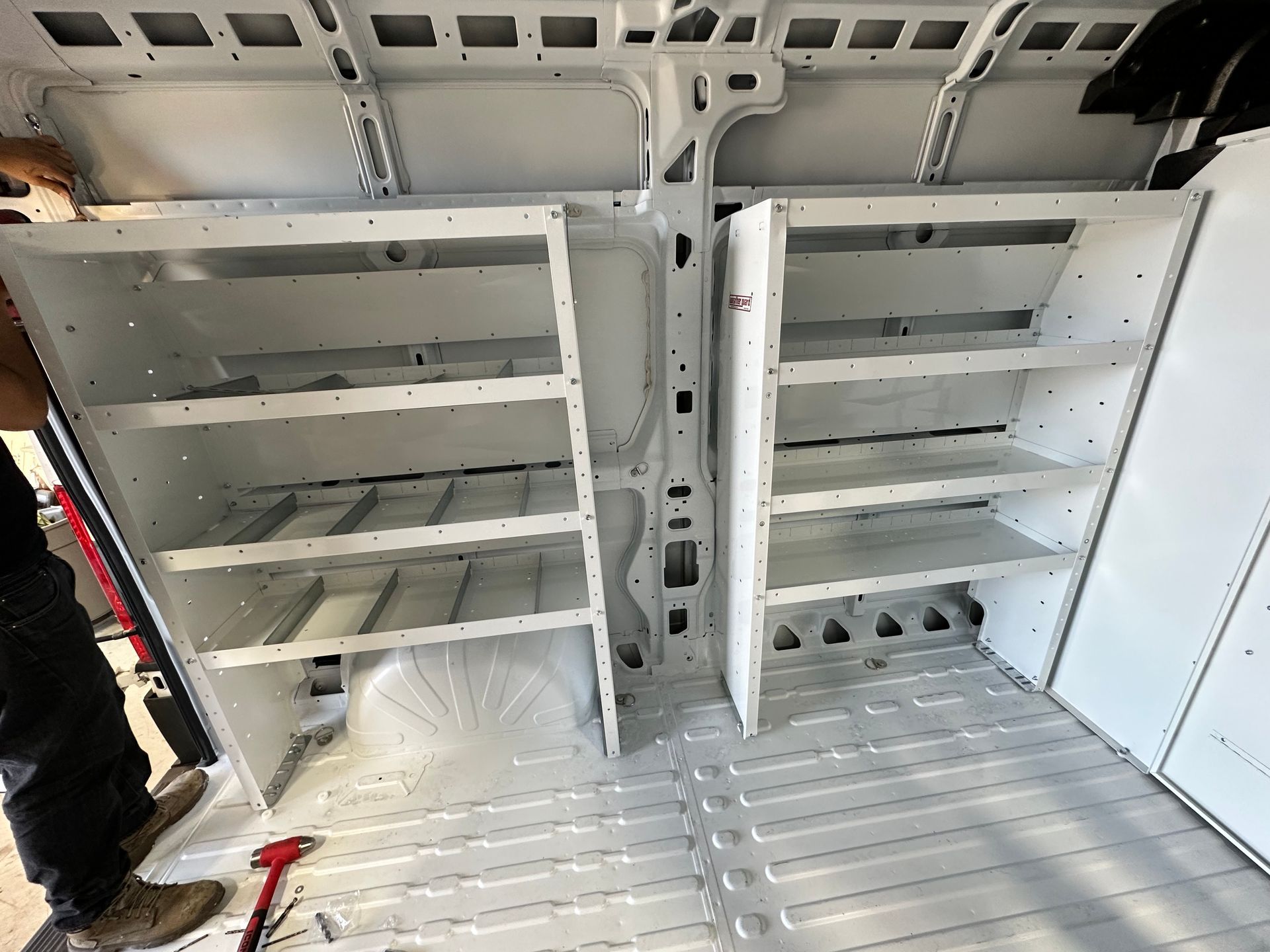 Two white metal shelving units installed inside the cargo area of a work van, viewed from the rear doors.