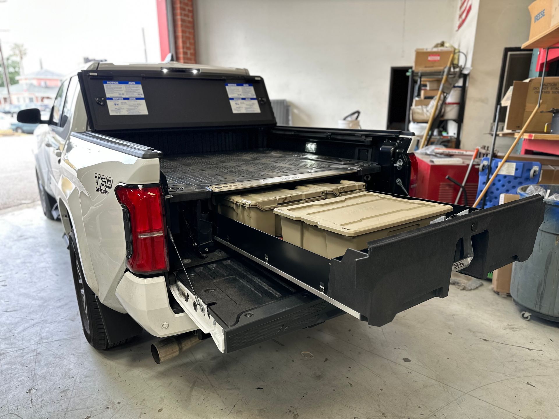 A white pickup truck with an open tailgate, revealing a black sliding cargo storage system holding tan plastic bins.