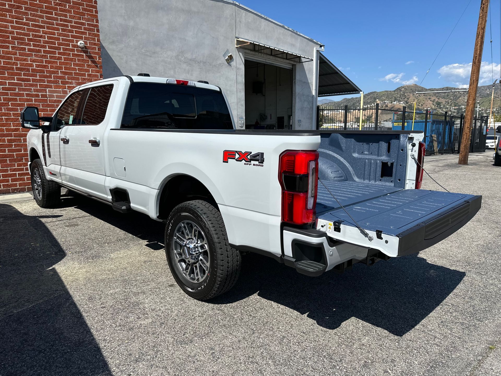 A white Ford Super Duty pickup truck with an open tailgate parked on a gravel lot next to a brick and concrete building.