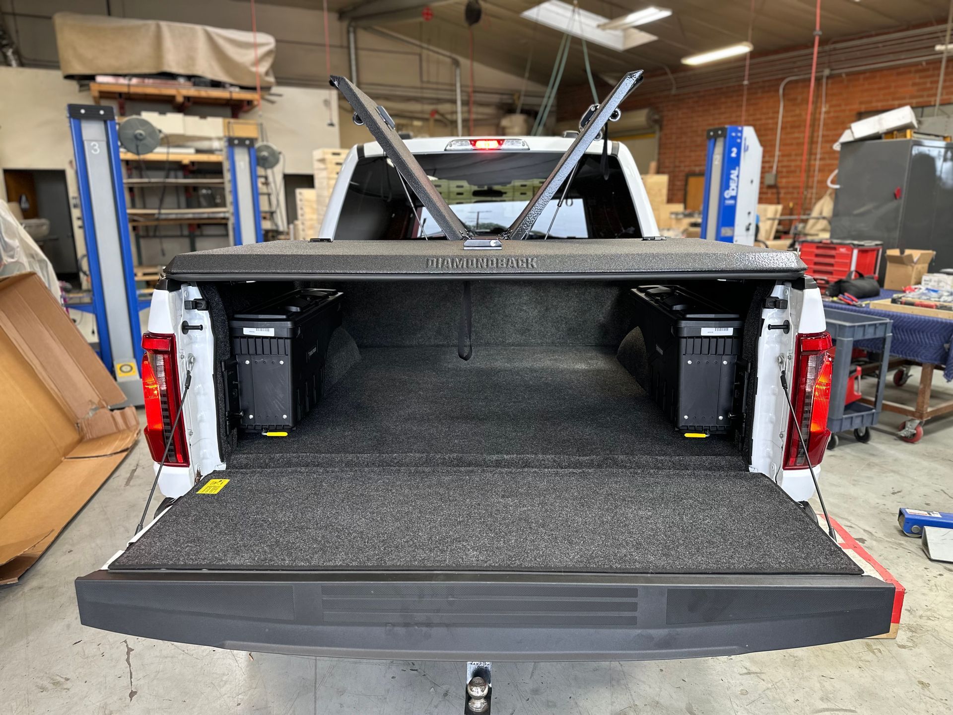 Open bed of a white pickup truck showing a black folding tonneau cover, grey carpeted liner, and two side storage boxes.