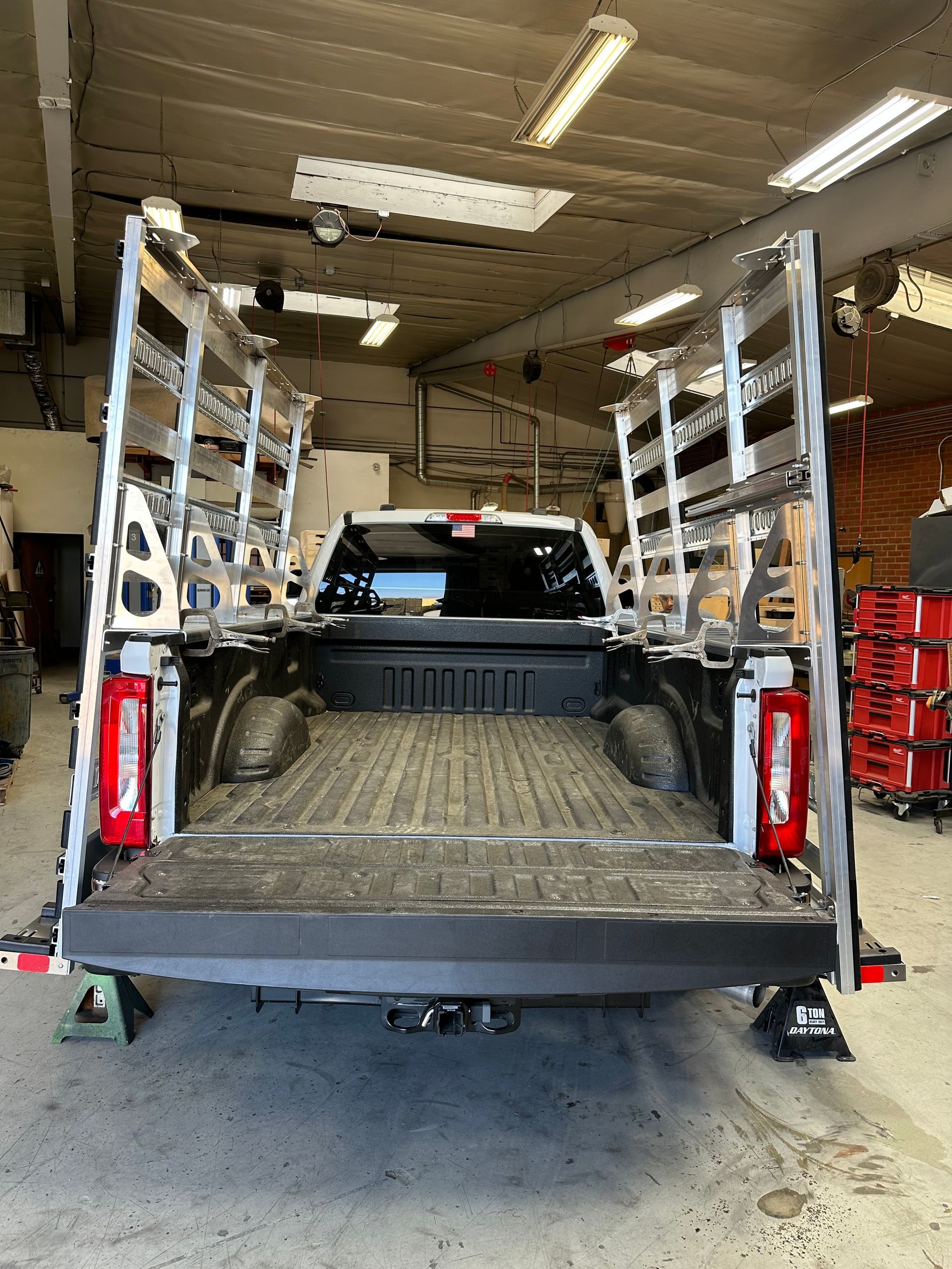 A white pickup truck parked in a garage, featuring tall, silver metal ladder racks mounted on both sides of the truck bed.