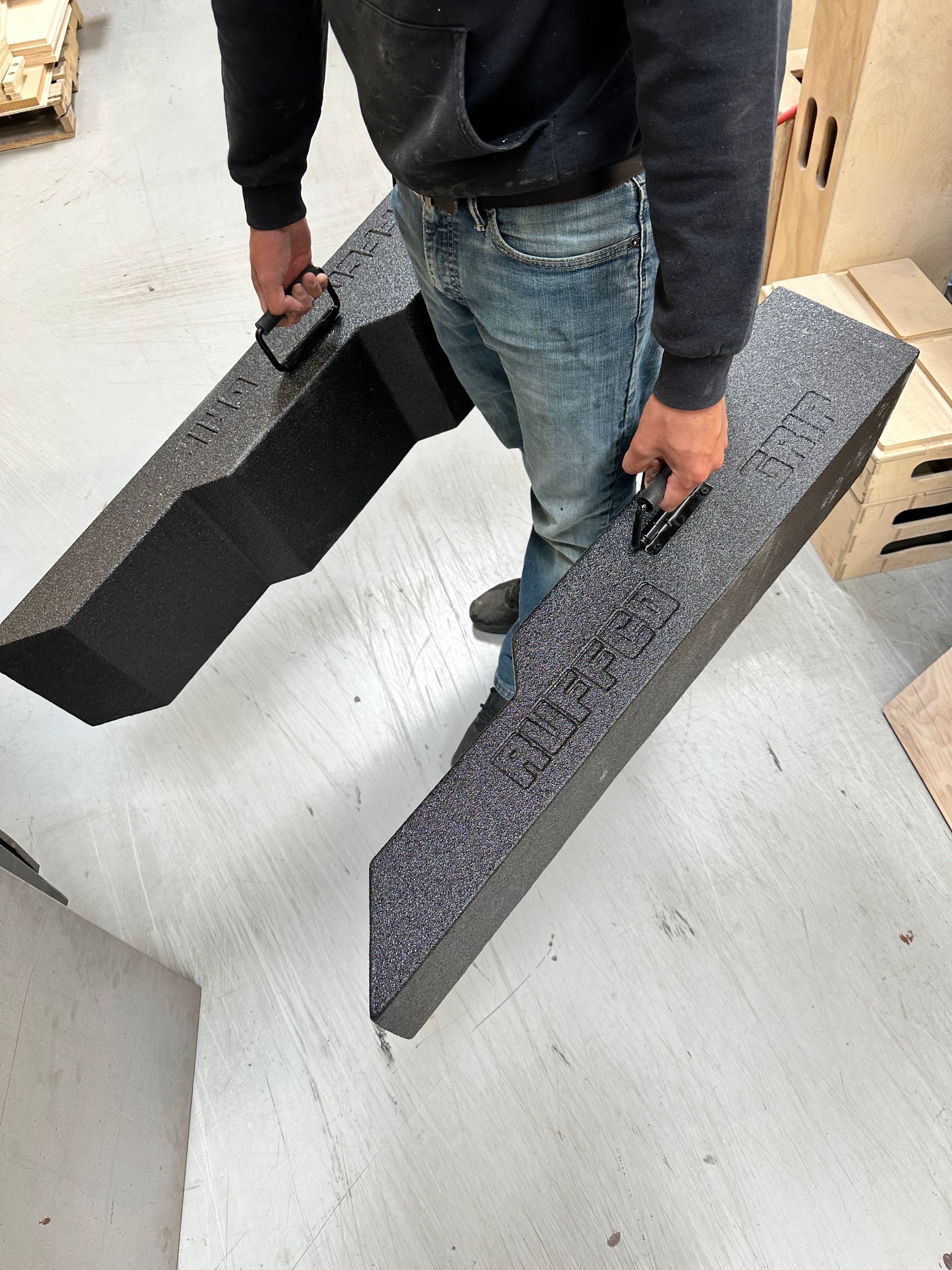 A person in a warehouse carries two long, dark, textured foam blocks, each equipped with a black handle.