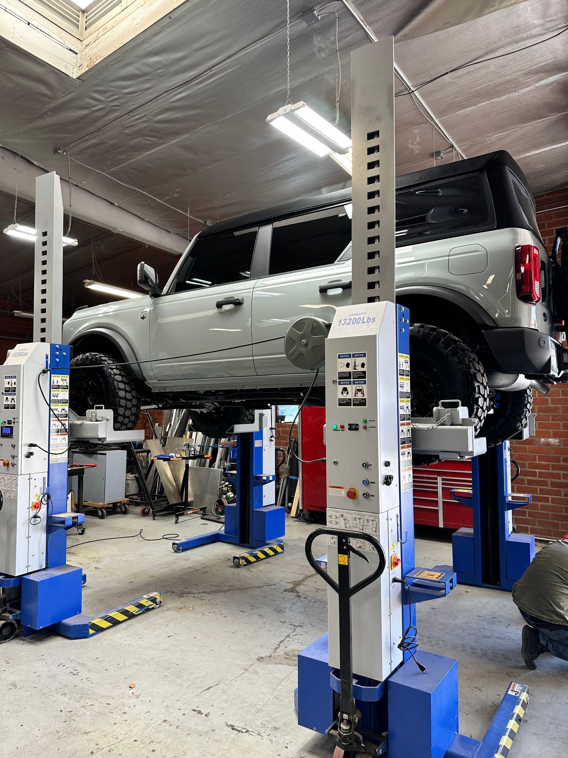 A light-colored SUV elevated on a mobile column lift system inside an automotive repair workshop.