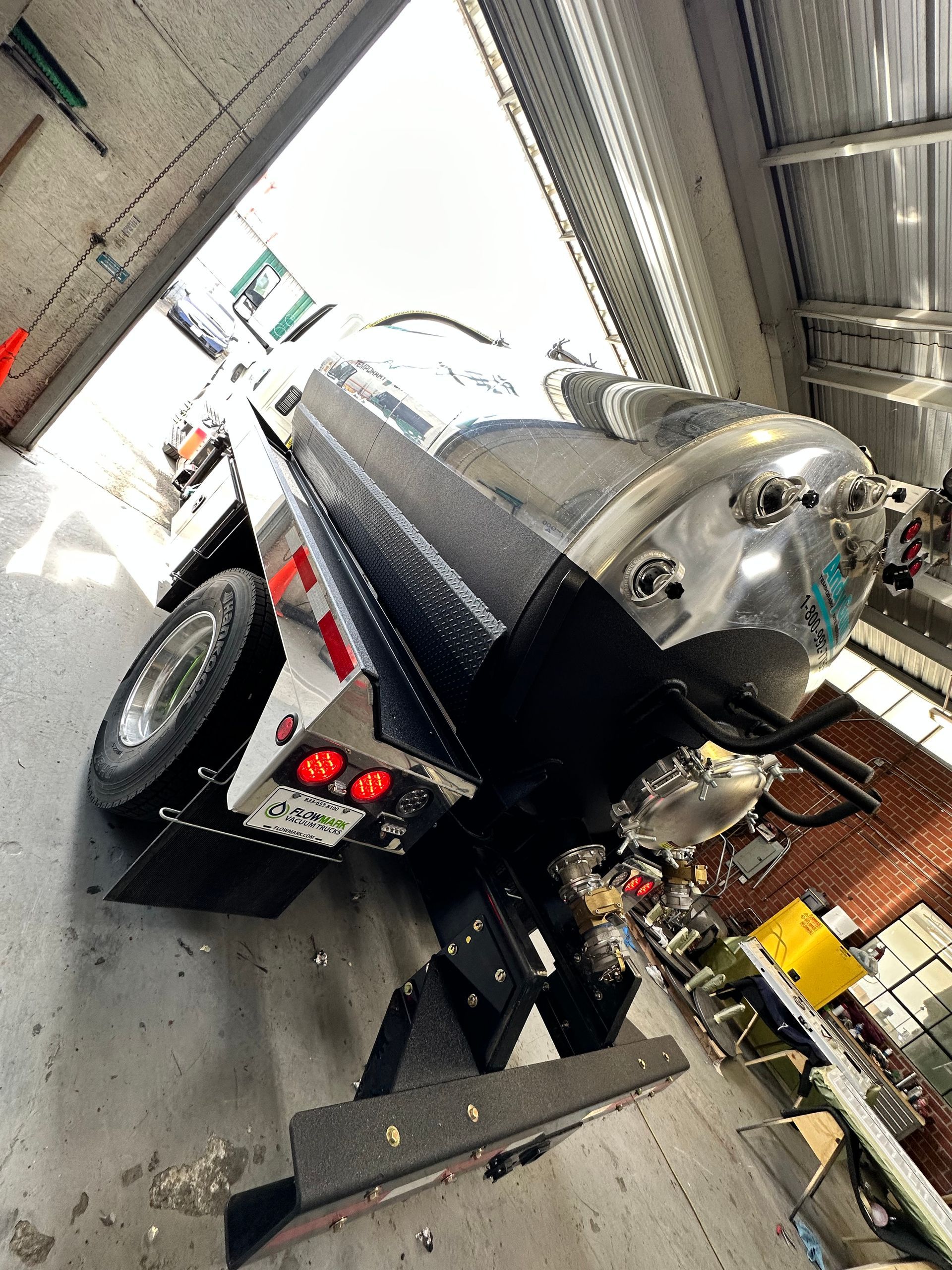 A stainless steel tanker trailer parked in a warehouse, showing its rear wheels, lighting, and plumbing valves.