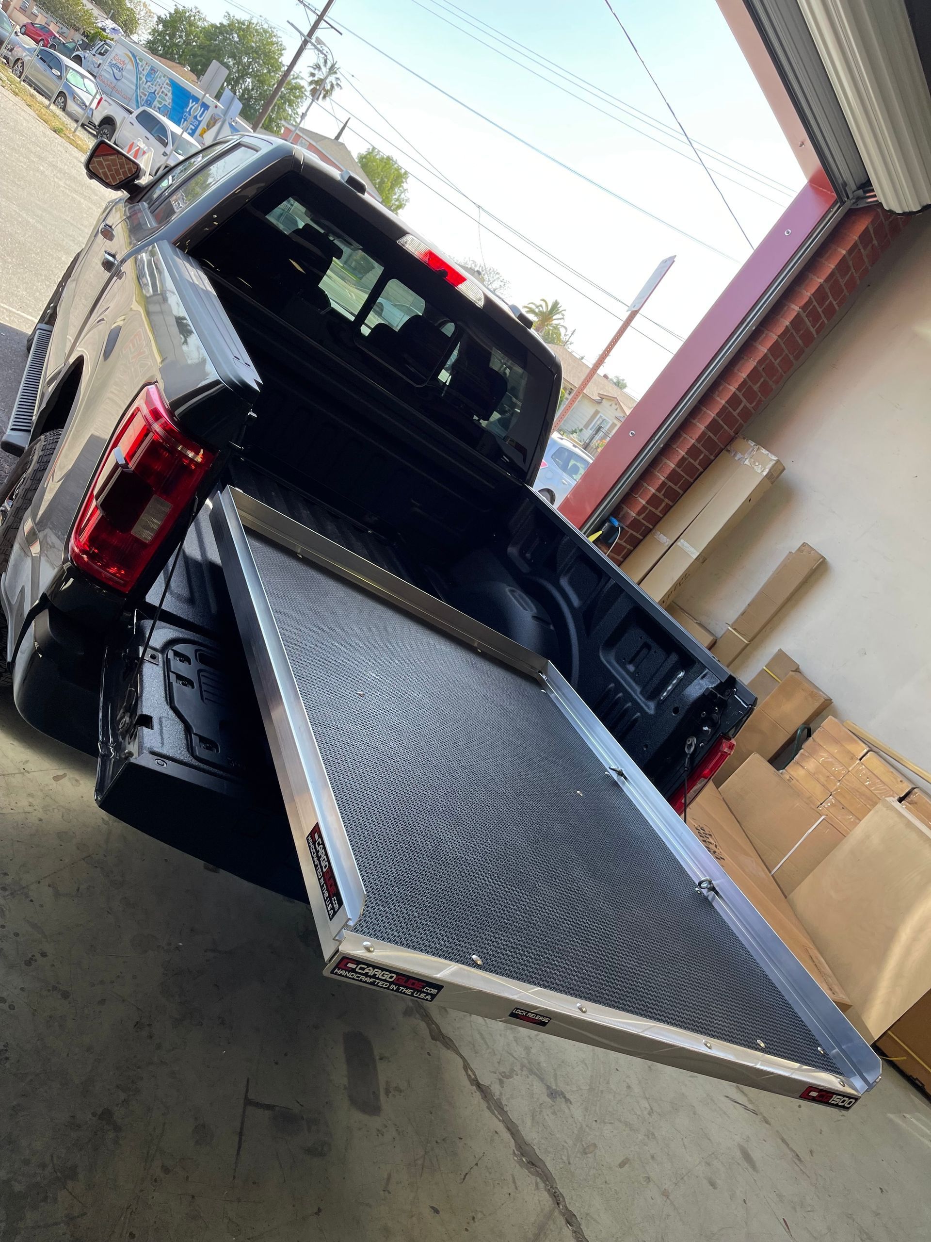 A black pickup truck bed with a metal sliding tray extended outward, sitting in an outdoor area near building materials.