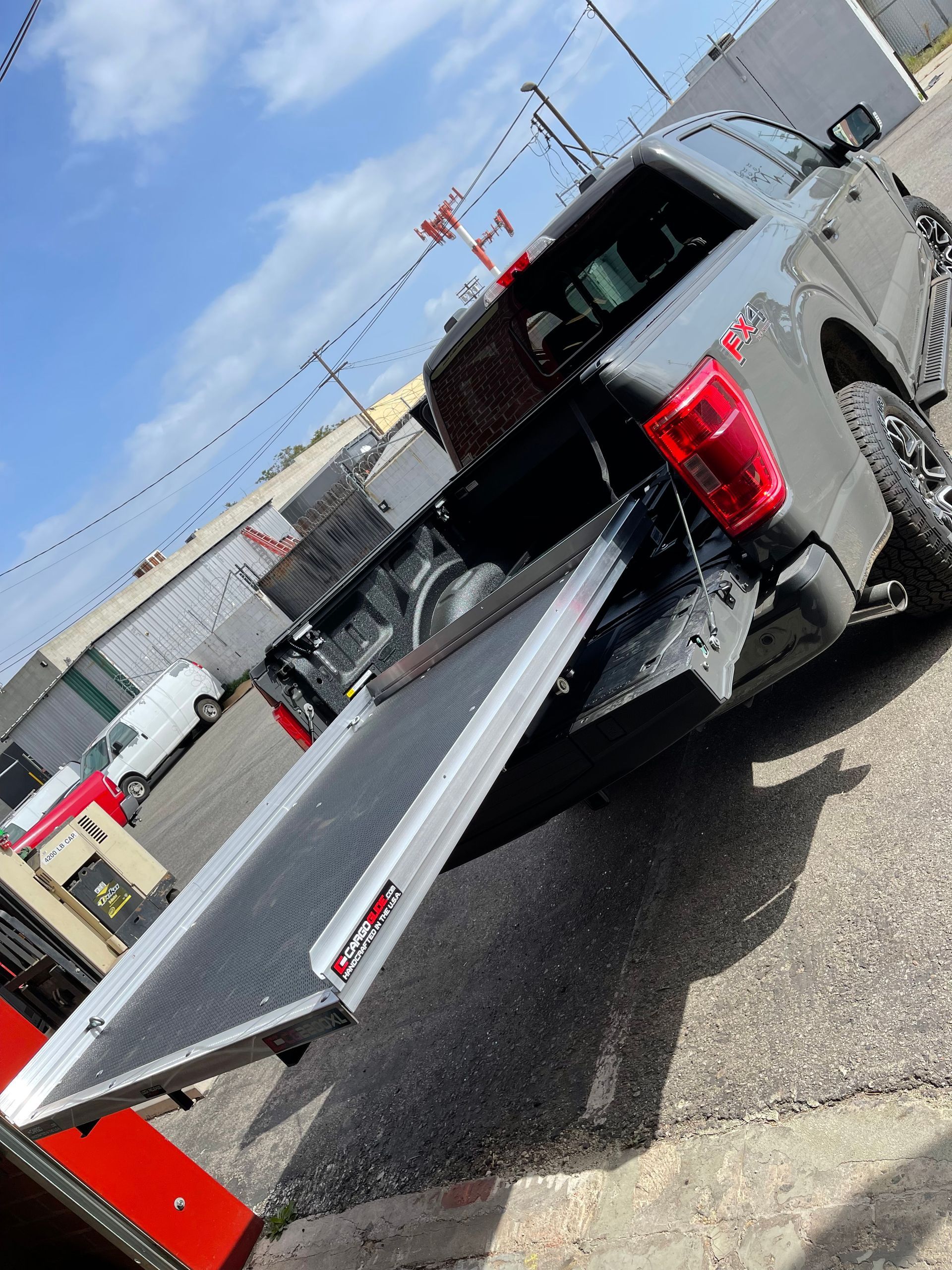A grey Ford F-150 pickup truck with an extended truck bed slide ramp deployed from its tailgate in a parking lot.