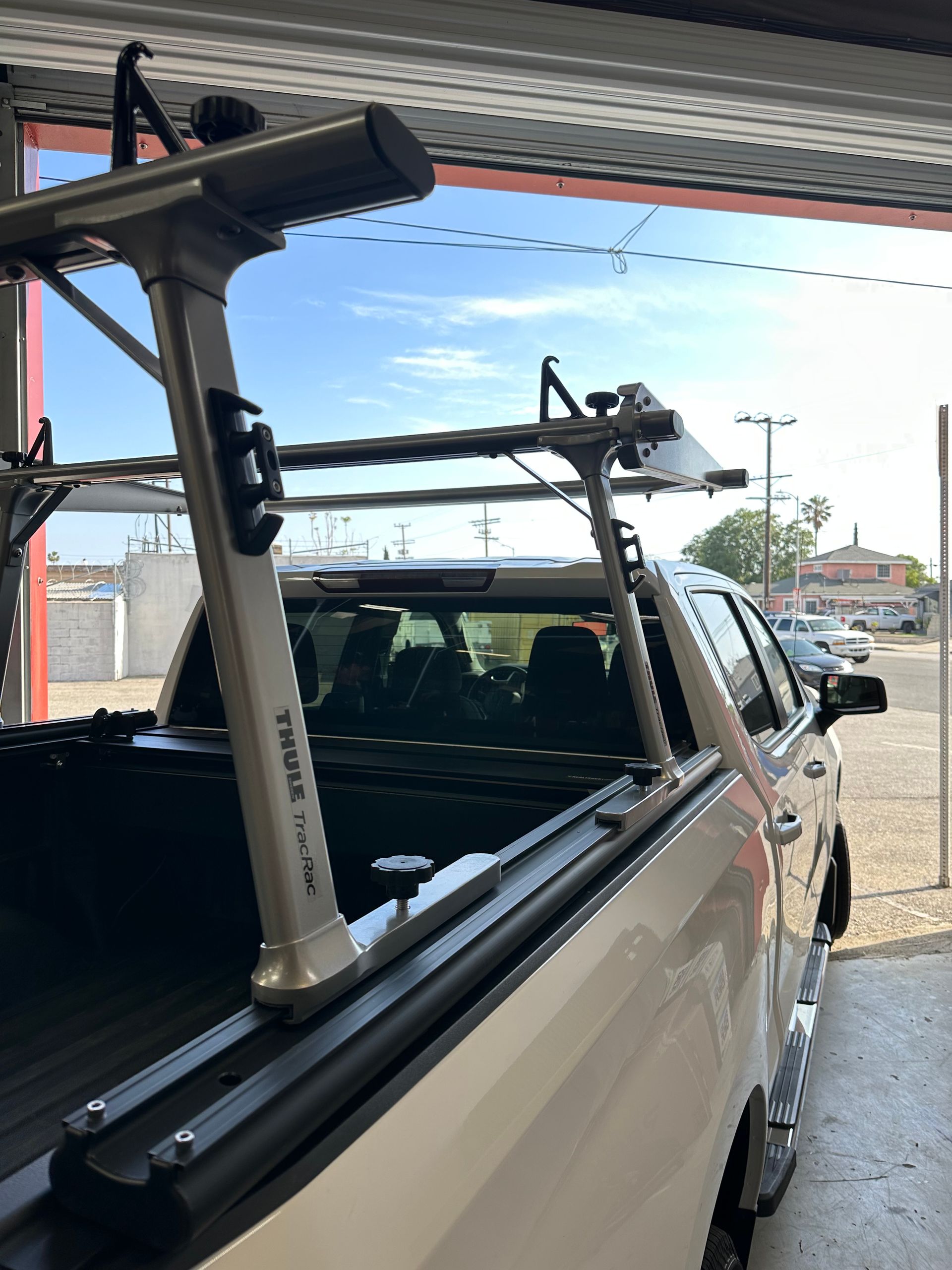 A silver truck bed rack installed on the side of a white pickup truck, seen from a high angle in a bright garage setting.