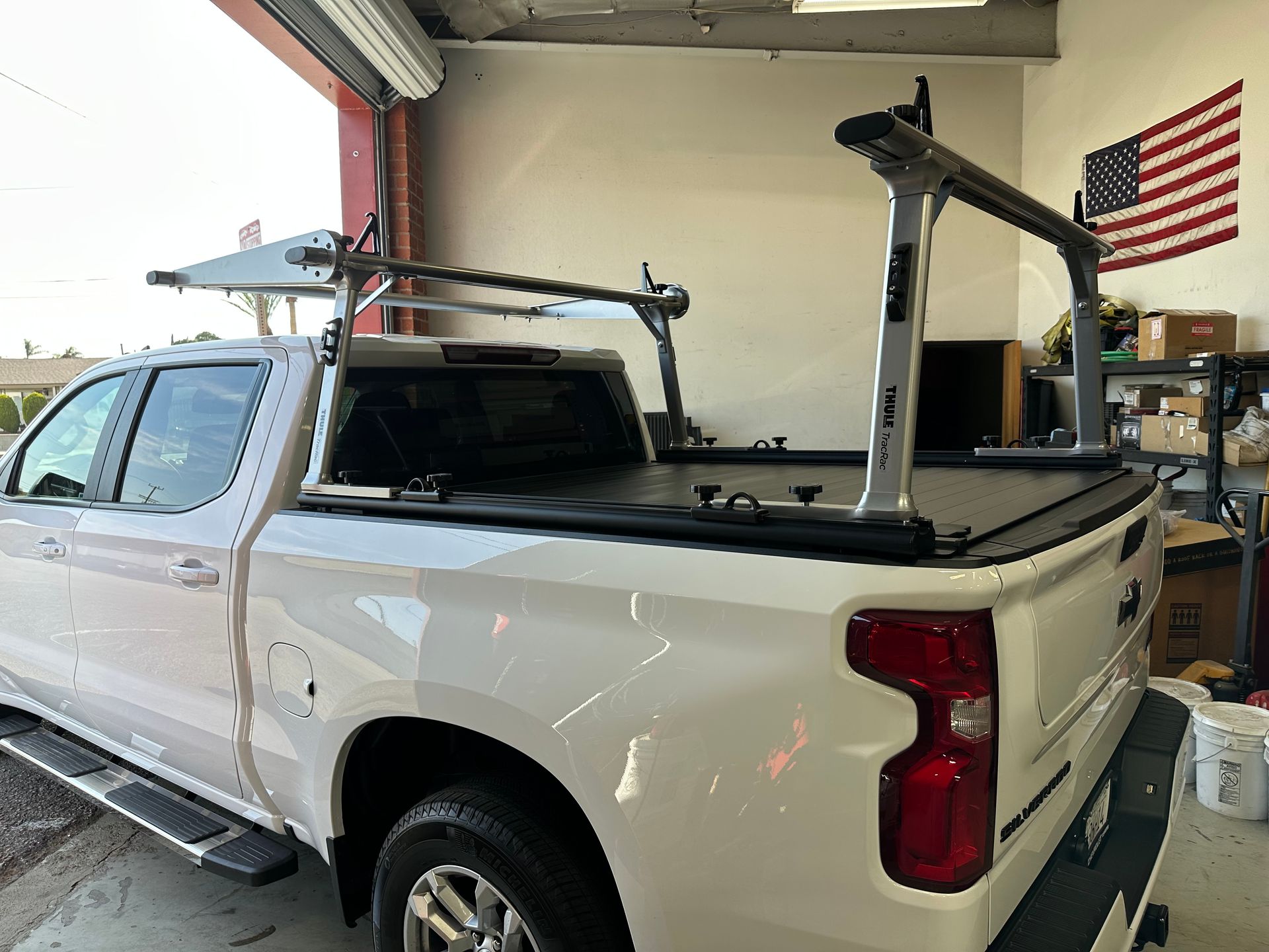 A white pickup truck parked in a garage, featuring a silver aftermarket metal ladder rack installed over the truck bed.