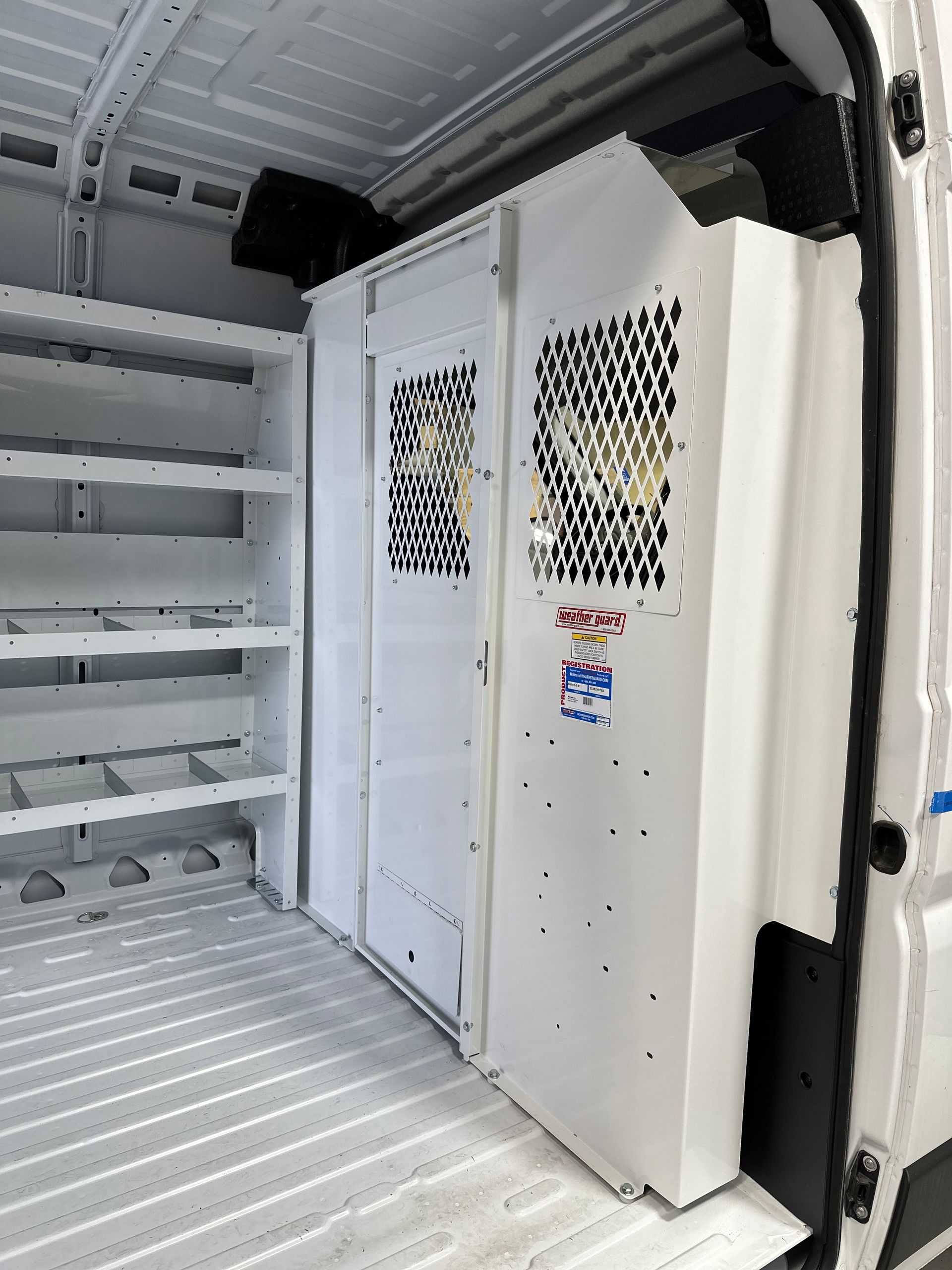 The interior of a white cargo van featuring a metal safety partition, a side door, and a white shelving unit.